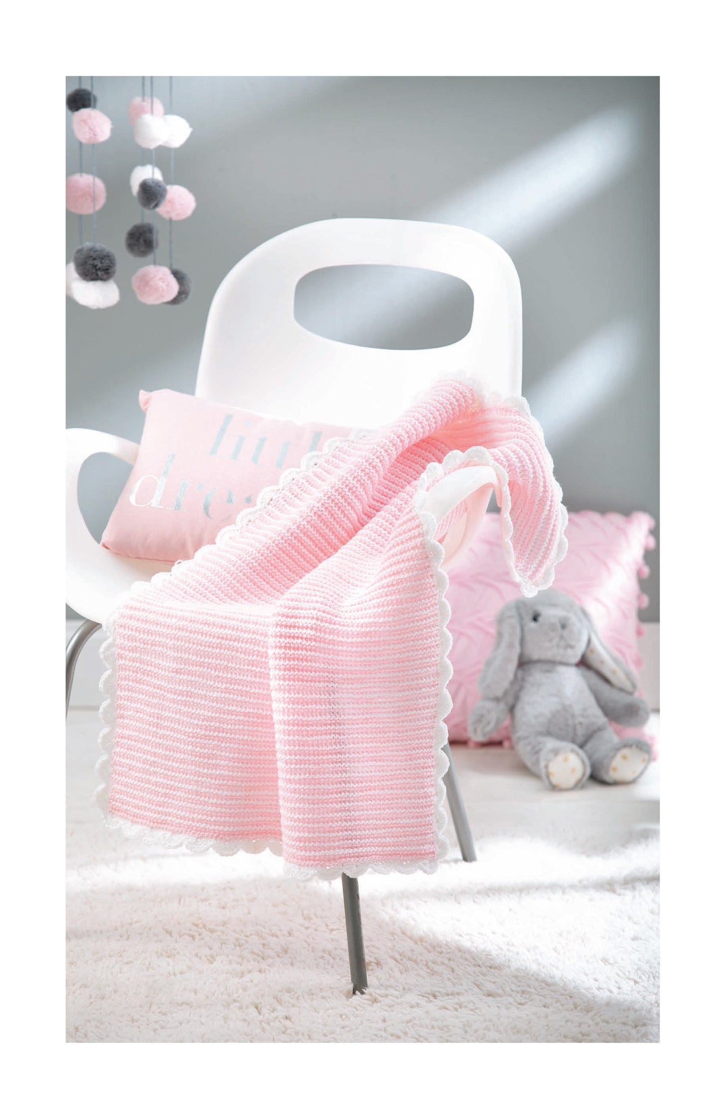 A cozy room features a white chair with a pink Tunisian crochet, pink pillow, plush gray bunny on a fuzzy rug, and pom-pom garland. Find inspiration in "Baby Bright Quilts" by Leisure Arts and use pops of color for stunning baby blanket designs!.