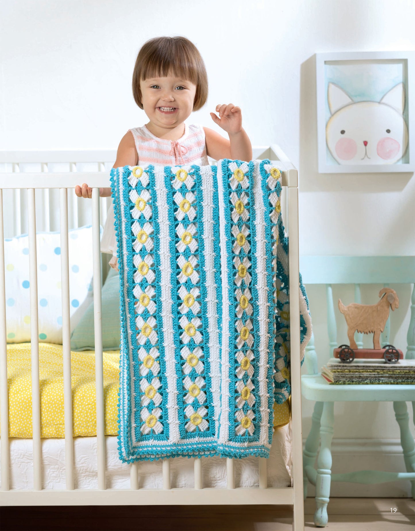 A smiling toddler stands in a white crib, holding the Leisure Arts Baby Bright Quilts—Use Pops of Color in Stunning Baby Designs! blanket, featuring blue, white, and yellow patterns. Playful animal art and a toy goat decorate the room.