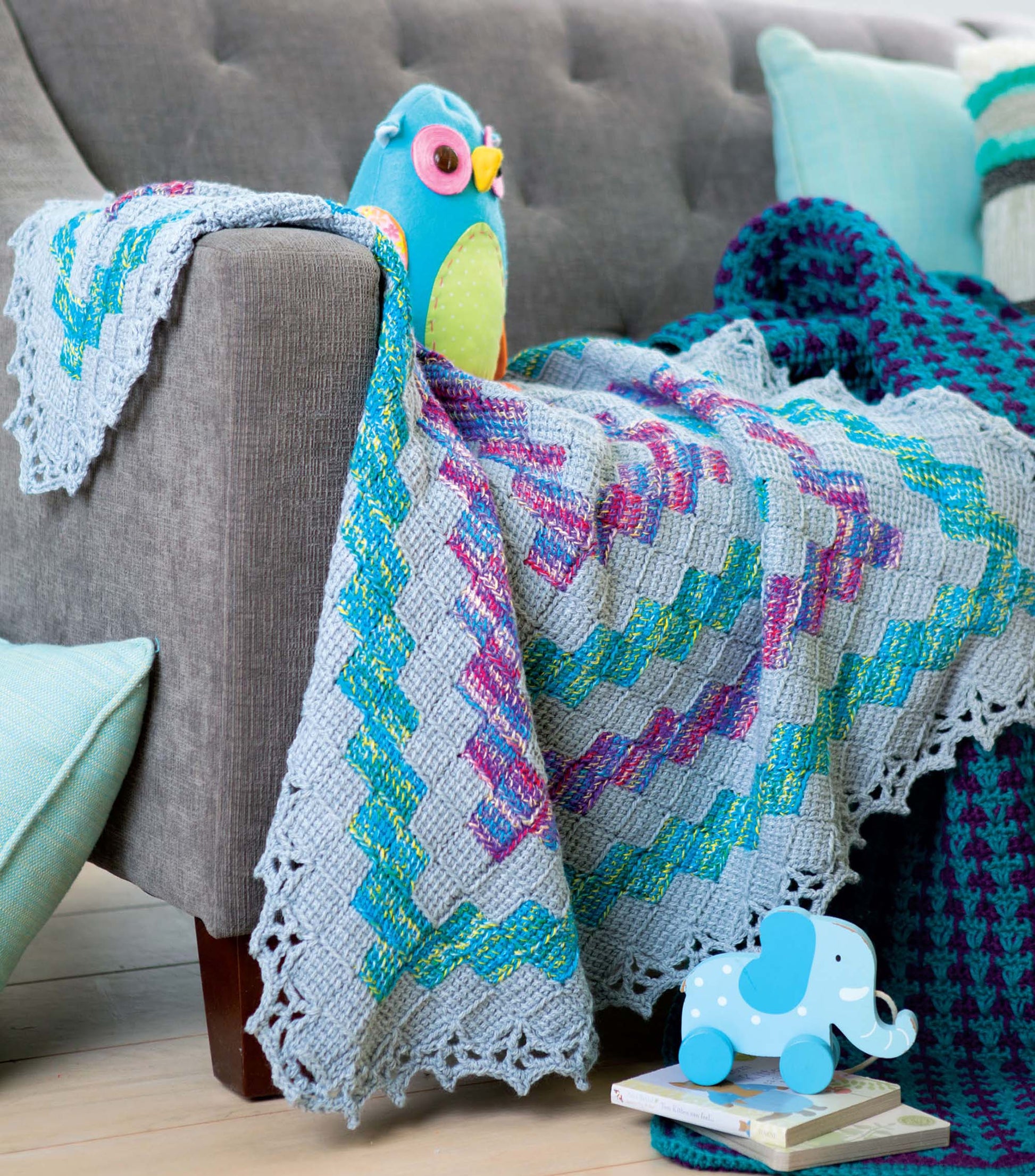 The "Afghans for the Family" book by Leisure Arts features colorful crochet blanket patterns with lace edges, shown draped over a gray sofa. A blue owl plush and toy elephant accent the scene, with pillows and another blanket in the background.