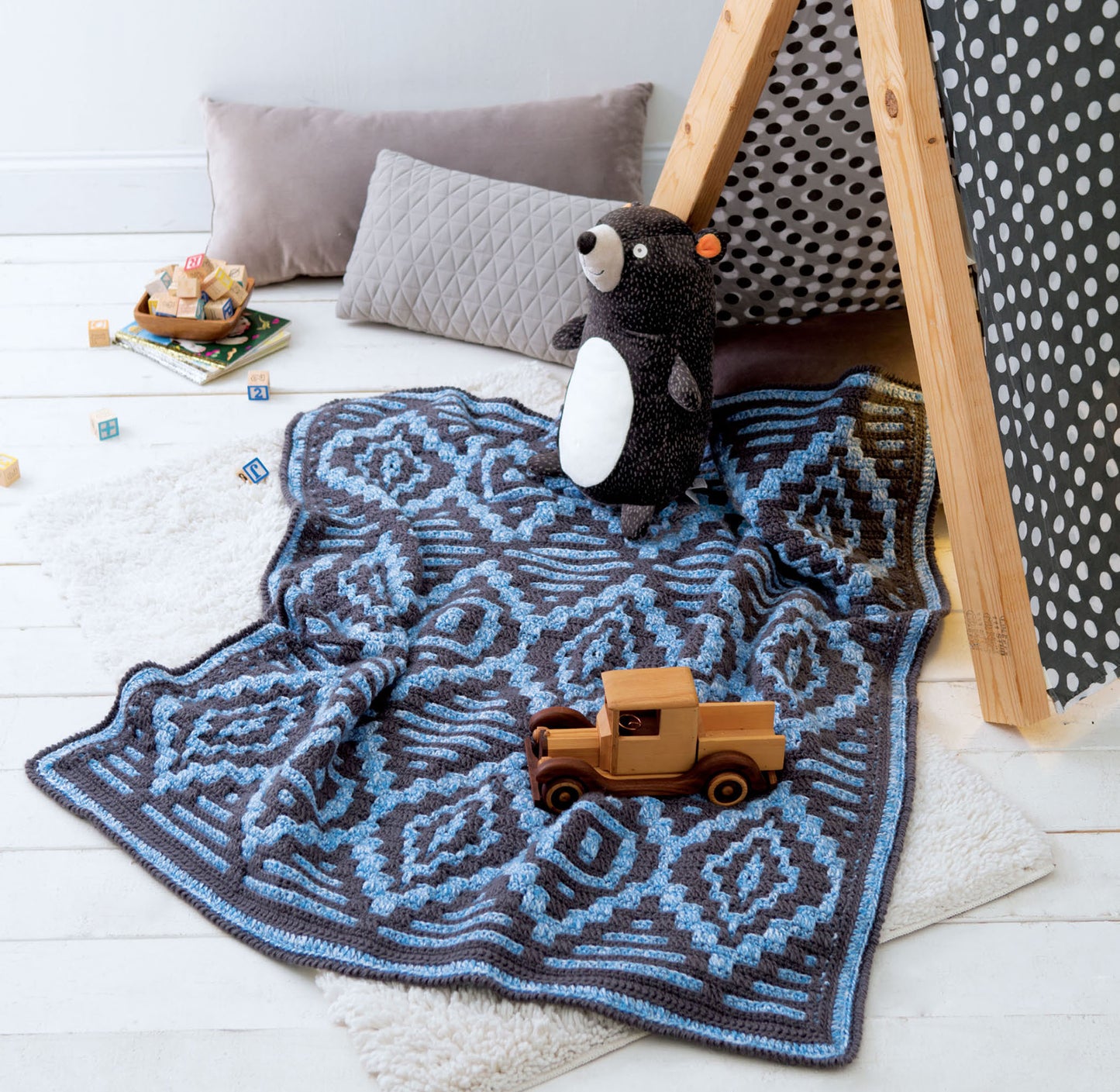 A cozy play area features Leisure Arts' "Afghans for the Family" blue and gray patterned blanket, a wooden toy truck, plush bear, gray pillows, and a wooden tent with polka dot fabric on a white floor.