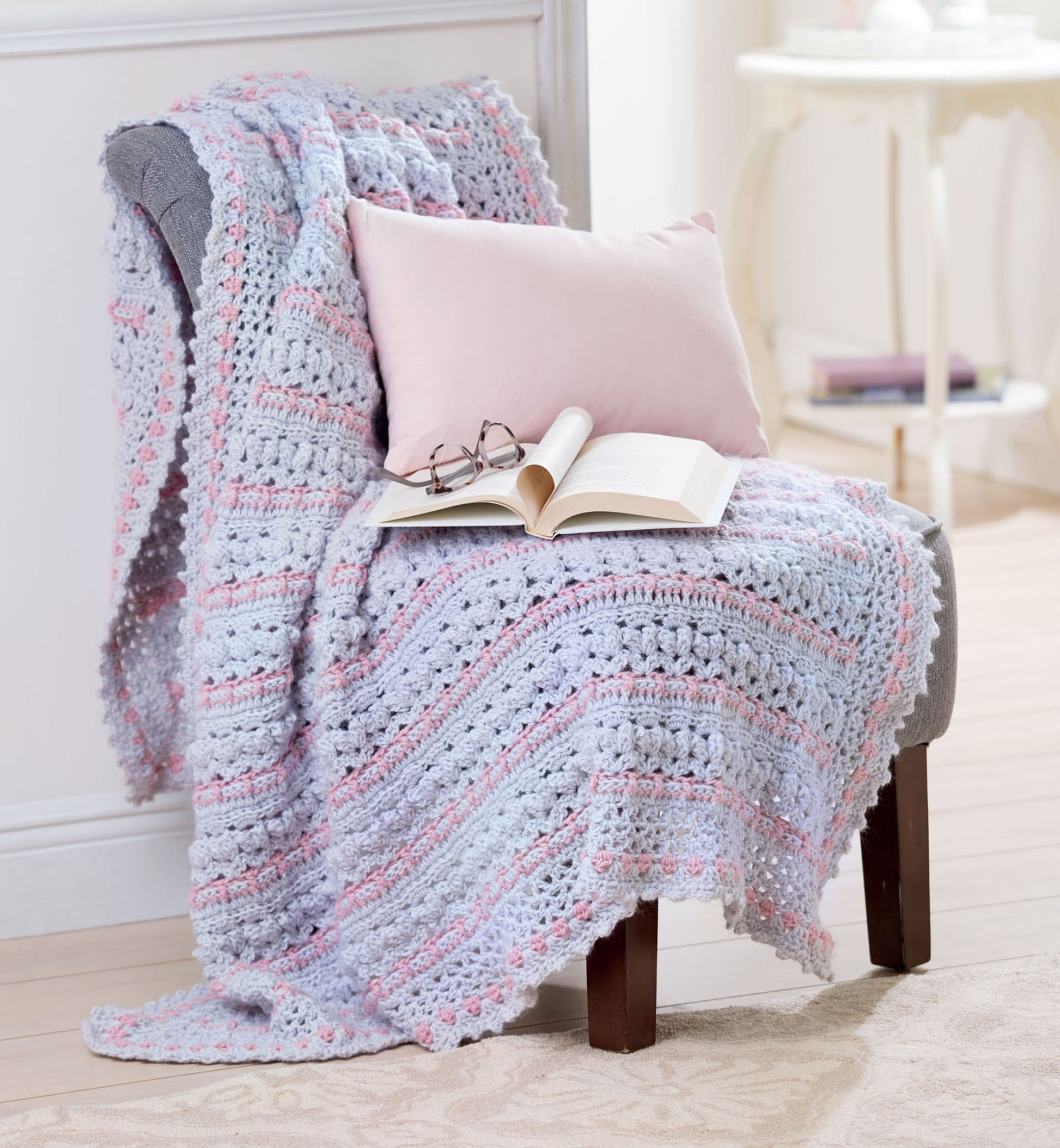A cozy armchair features a Leisure Arts "Afghans for the Family" pastel crochet blanket and a pale pink pillow, with an open book and reading glasses on the blanket, in a bright, inviting room.