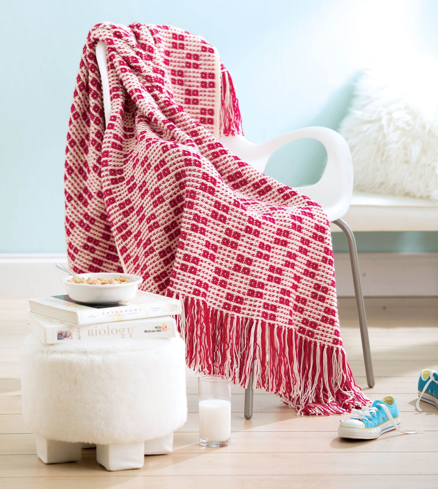 The "Afghans for the Family" by Leisure Arts—a red and white crochet-patterned throw—rests on a modern white chair, with a furry ottoman, books, cereal bowl, milk glass, and blue sneakers on the light wood floor nearby.