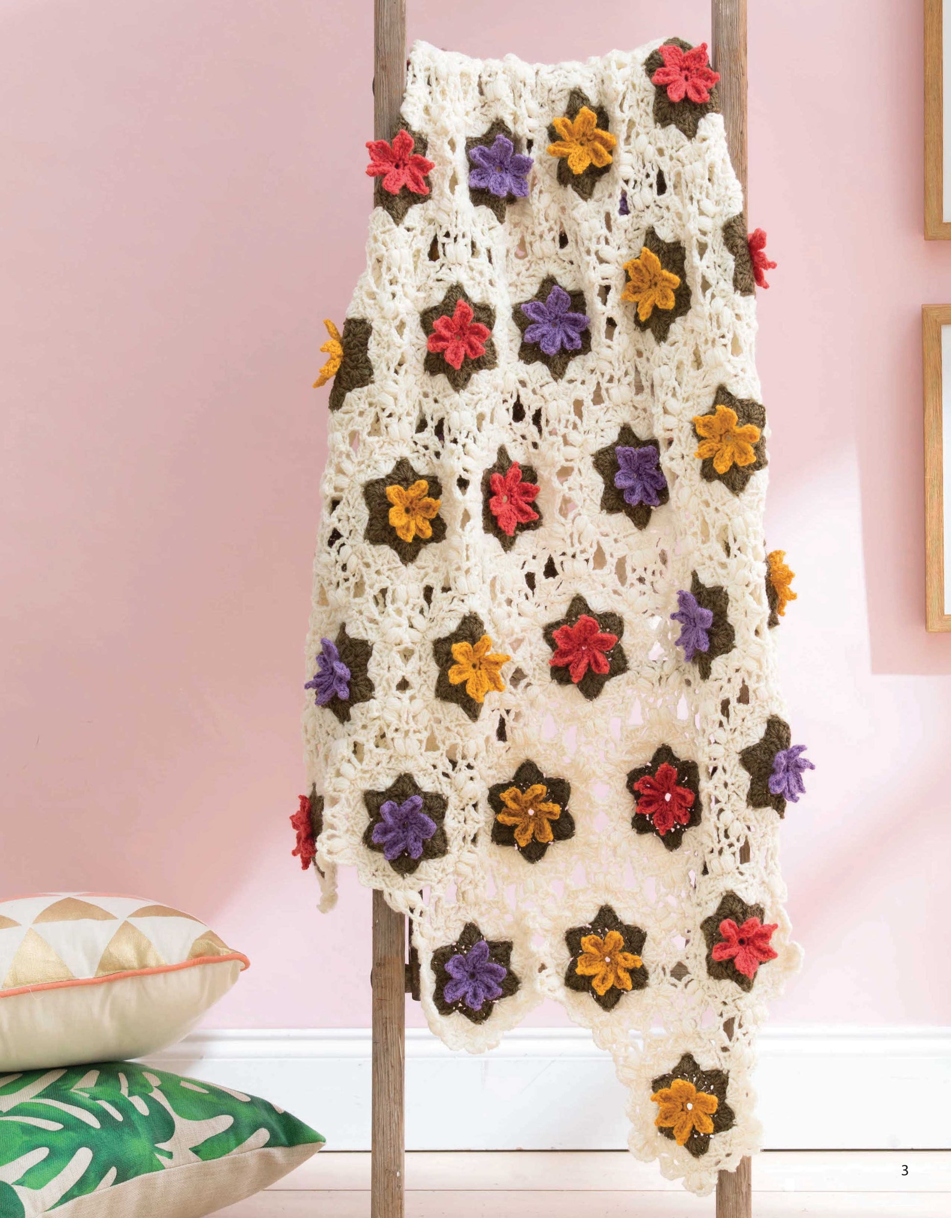 The "Grannies In Bloom Digital Download" by Leisure Arts is a cream-colored crochet blanket with 3D granny motifs in red, yellow, orange, and purple, displayed over a wooden ladder above a pillow with green leaf patterns against a pink wall.