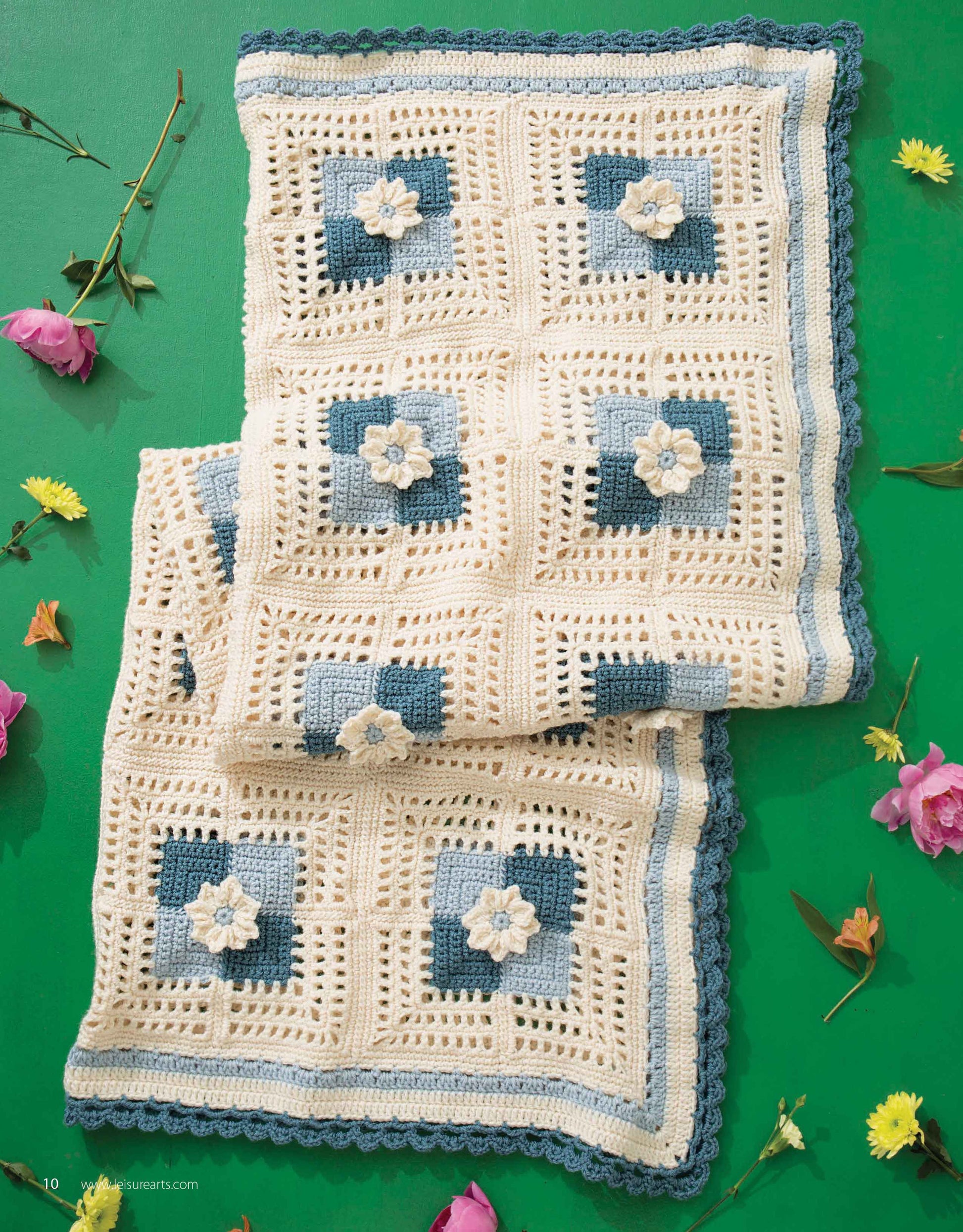 The "Grannies In Bloom Digital Download" by Leisure Arts showcases a crocheted blanket with cream, blue, and navy granny flower motifs, displayed on a green background adorned with pink and yellow flowers.