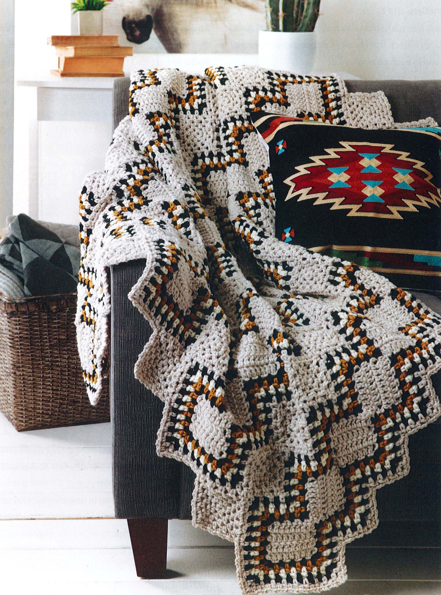 The Modern Southwest Afghans by Leisure Arts, featuring geometric crochet in beige, black, yellow, and brown, is draped over a gray sofa with a vibrant Southwestern cushion and a nearby basket. A potted cactus adds a touch of greenery.