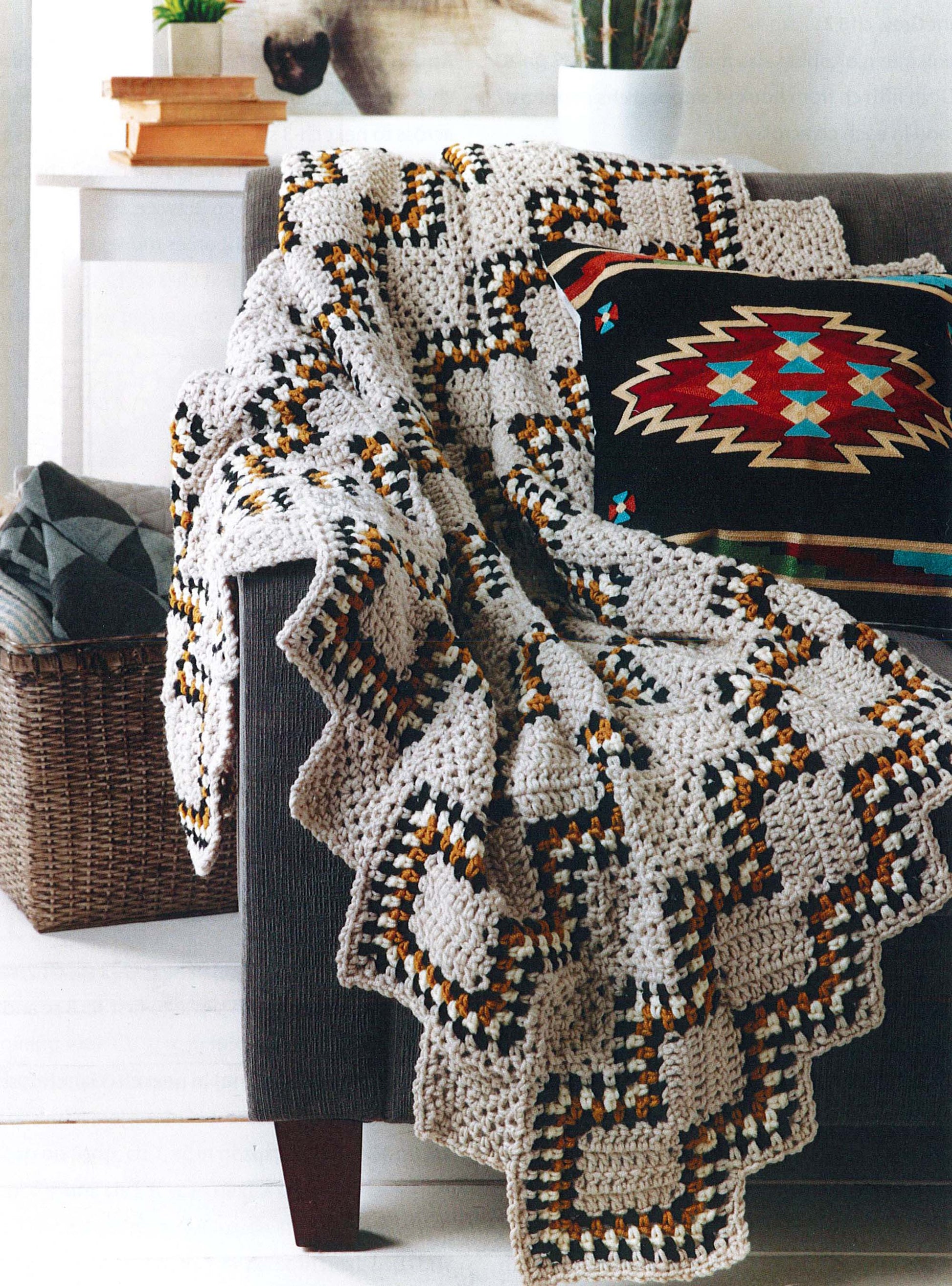 The Modern Southwest Afghans by Leisure Arts, featuring geometric crochet in beige, black, yellow, and brown, is draped over a gray sofa with a vibrant Southwestern cushion and a nearby basket. A potted cactus adds a touch of greenery.