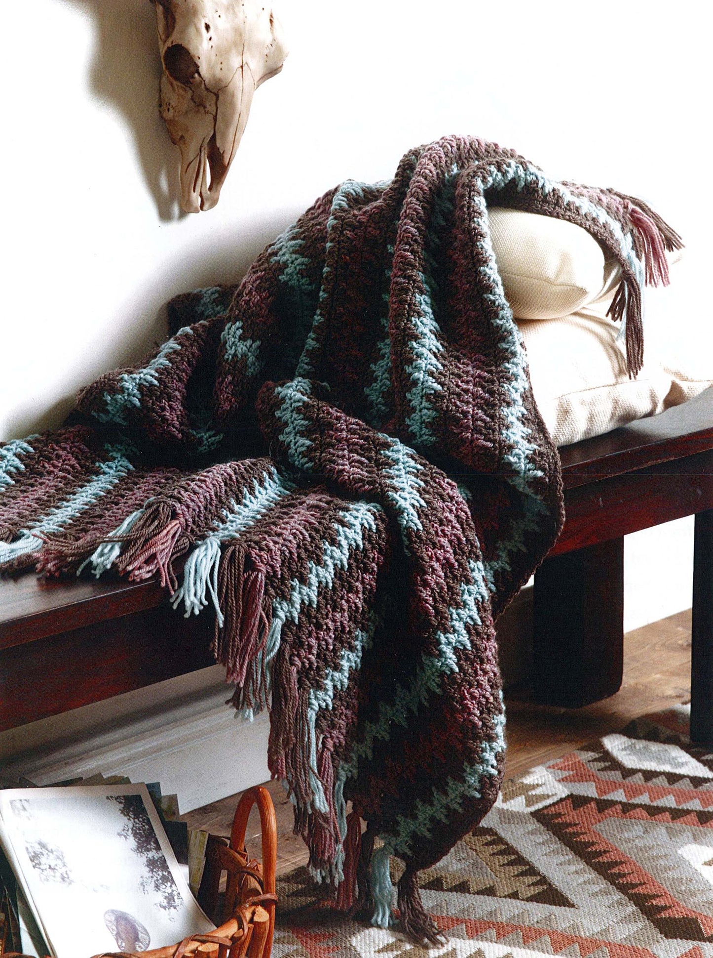 The Modern Southwest Afghans by Leisure Arts, featuring blue and brown stripes with fringe edges, is draped over a cushioned wooden bench. A patterned rug and magazines lie on the floor, with a skull accent on the white wall above.