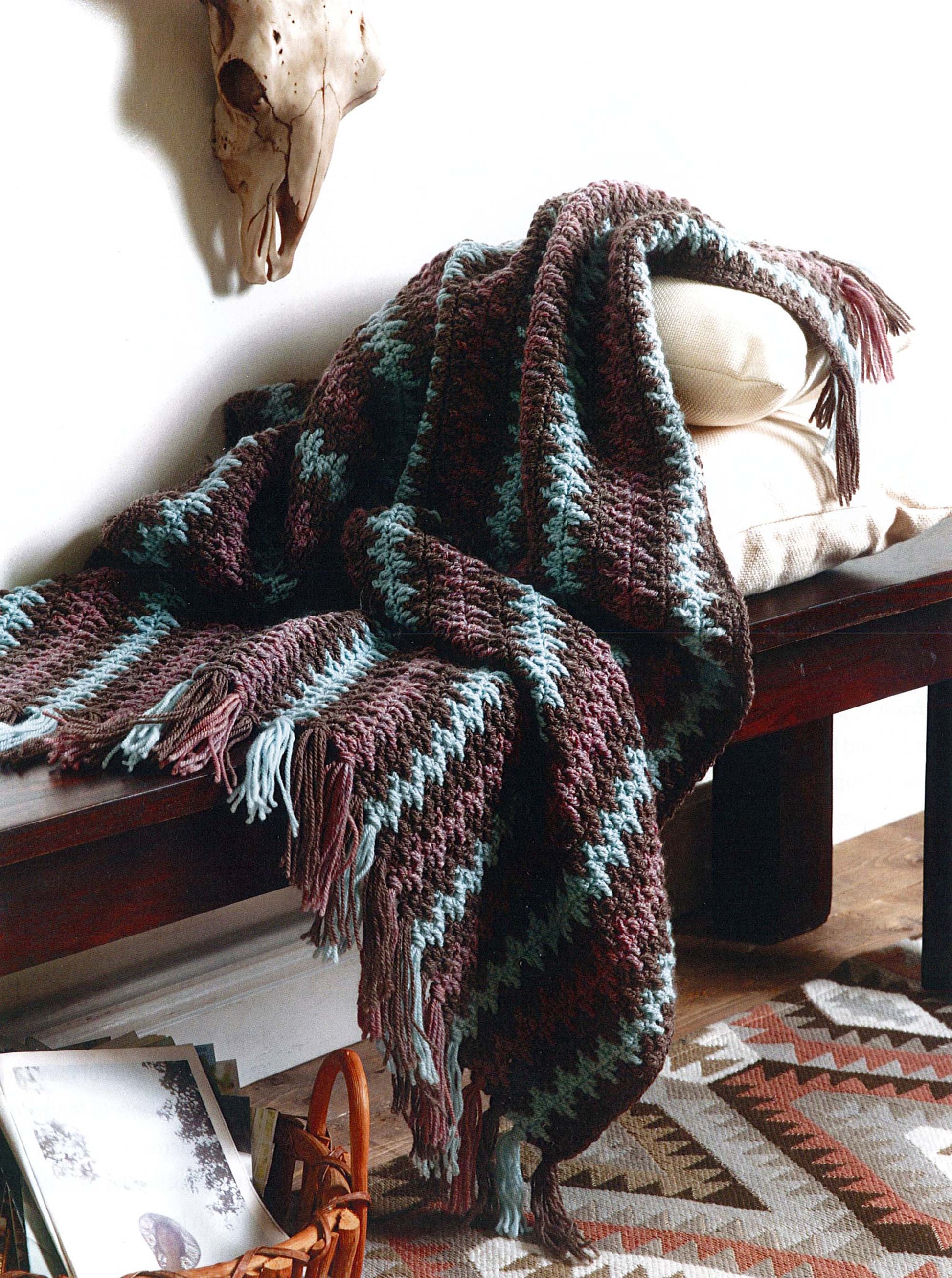 The Modern Southwest Afghans by Leisure Arts, featuring blue and brown stripes with fringe edges, is draped over a cushioned wooden bench. A patterned rug and magazines lie on the floor, with a skull accent on the white wall above.