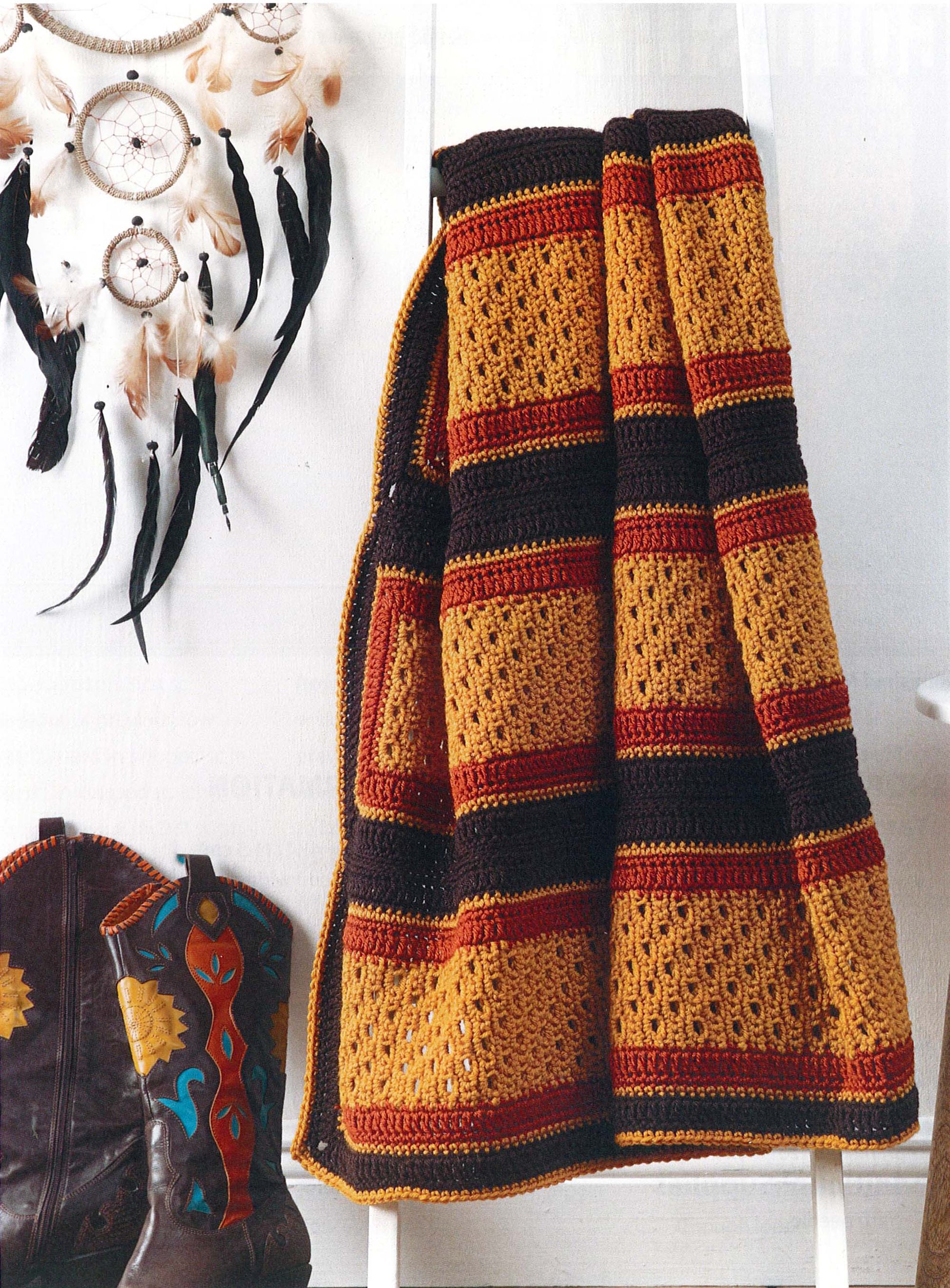 A Leisure Arts "Modern Southwest Afghans" blanket with mustard yellow, rust red, and dark brown stripes hangs on a white ladder beside embroidered cowboy boots and a wall of feathered dreamcatchers.