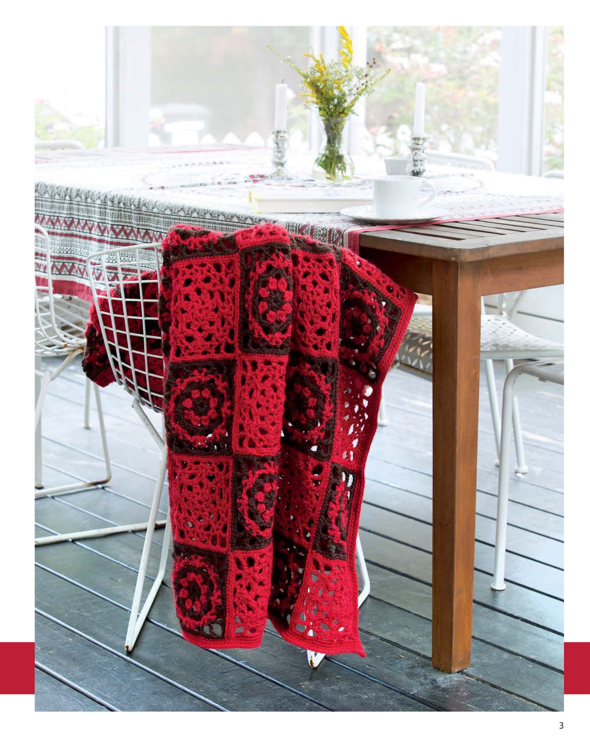 A cozy crocheted blanket from "Blocks and Stripes—7 Cozy Projects Using Easy Granny Squares & Strips" by Leisure Arts is draped over a white wire chair beside a floral-covered wooden table with candlesticks and yellow flowers on the porch.
