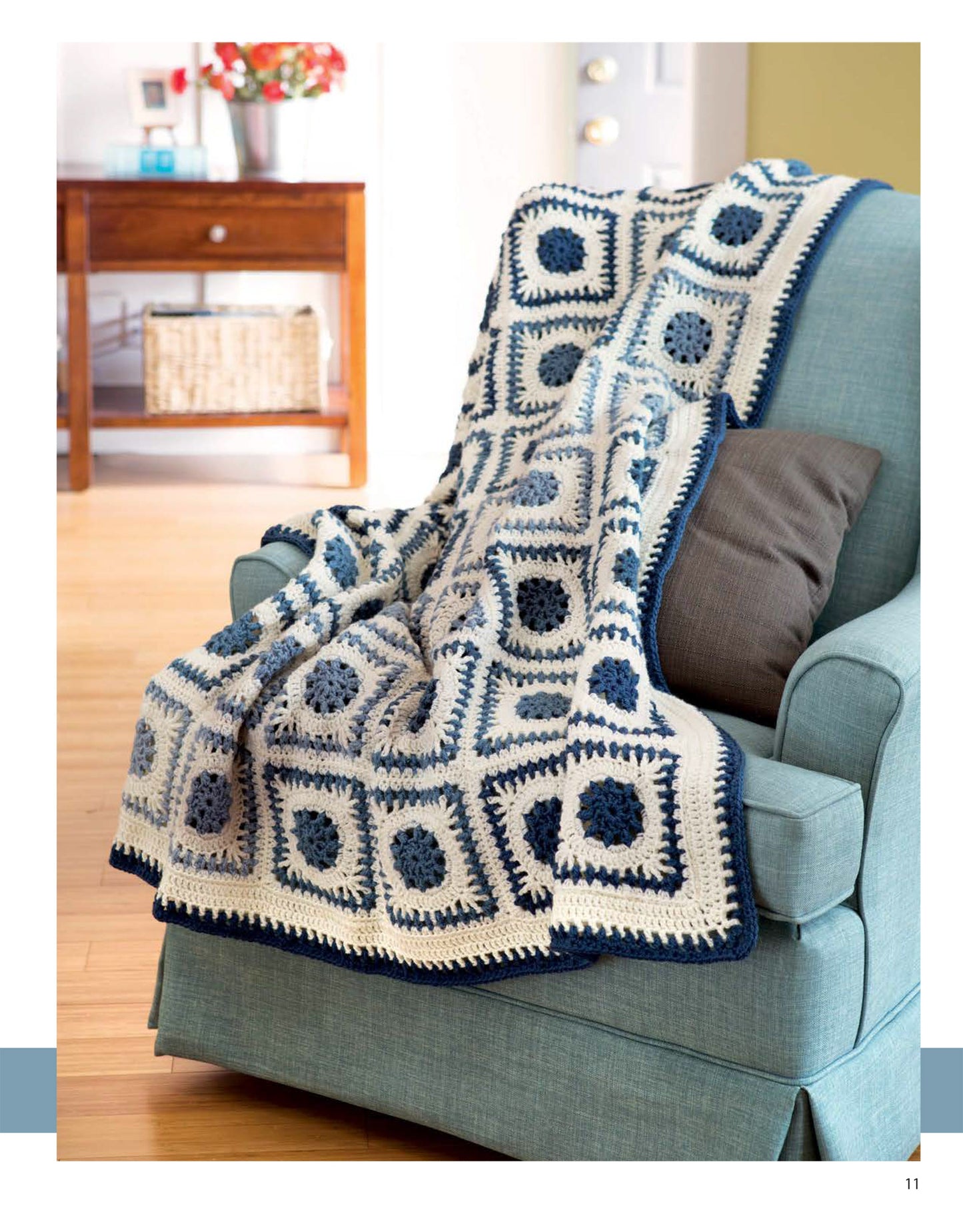 The "Blocks and Stripes—7 Cozy Projects Using Easy Granny Squares & Strips" by Leisure Arts features a blue and white crochet blanket with classic square motifs, draped over a light blue armchair in a bright, inviting room.