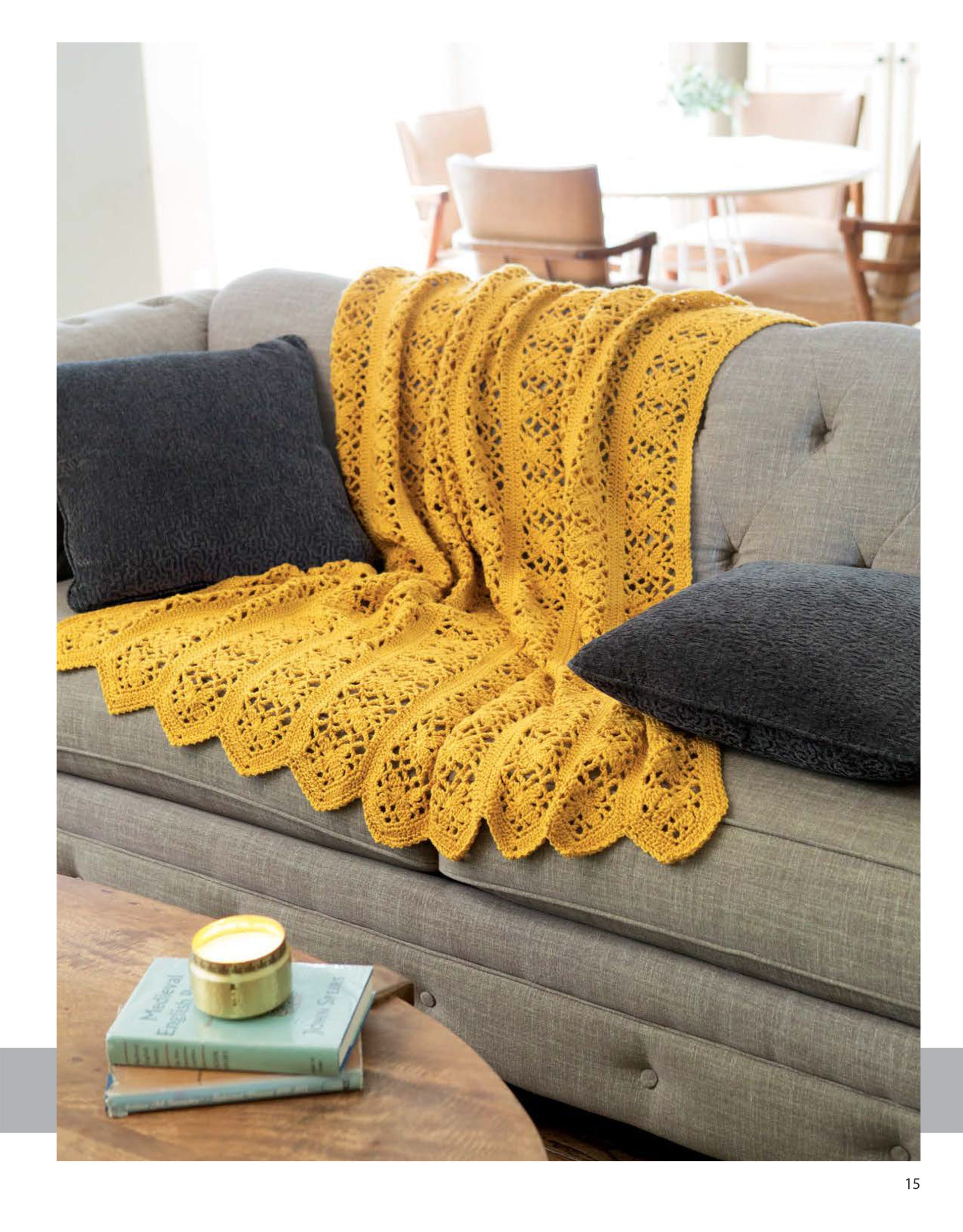 A cozy crochet blanket from Leisure Arts' "Blocks and Stripes—7 Cozy Projects Using Easy Granny Squares & Strips" is draped over a light gray sofa with two dark gray pillows, near a round wooden coffee table and dining set in the background.