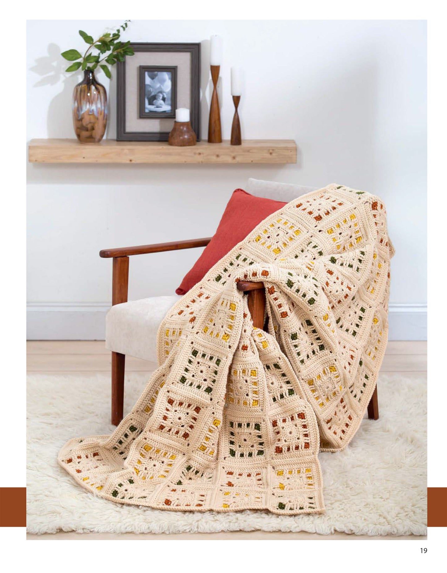 A cream armchair with a coral cushion sits on a white rug. Draped over it is "Blocks and Stripes—7 Cozy Projects Using Easy Granny Squares & Strips" by Leisure Arts. Behind, a wooden shelf holds decor and a framed photo.