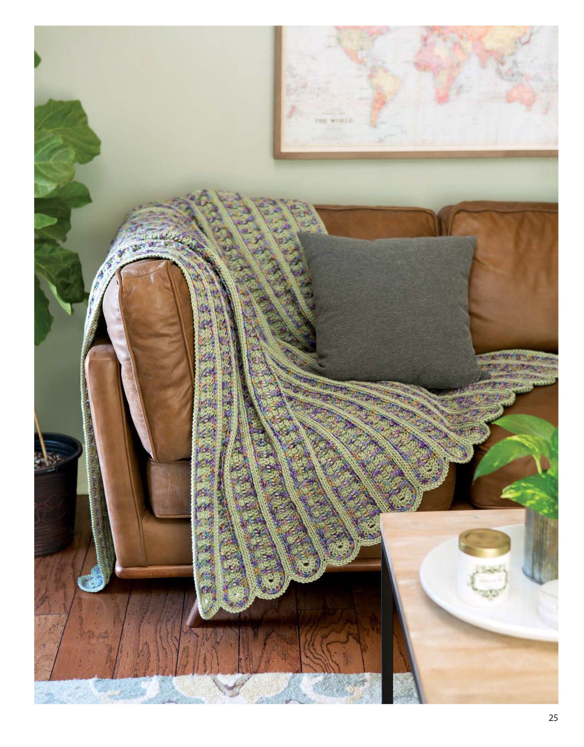 A tan leather sofa with a gray pillow features a cozy blanket inspired by Blocks and Stripes—7 Cozy Projects Using Easy Granny Squares & Strips from Leisure Arts. A world map, potted plant, and candles accent the inviting space.