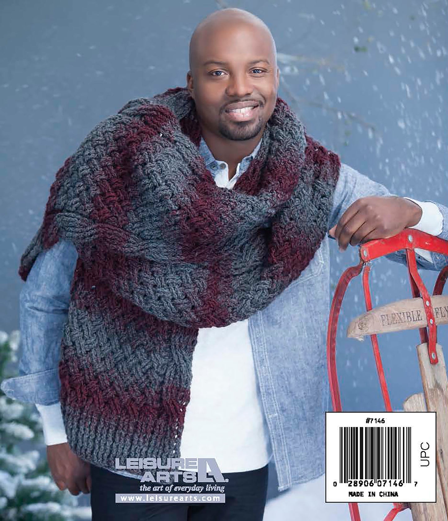 A man smiles outdoors in the snow, wearing a chunky maroon and gray scarf made with Leisure Arts’ Easy Super Scarves Crochet, perfect for beginners. He leans on a vintage sled, dressed in a light blue shirt and brown pants.