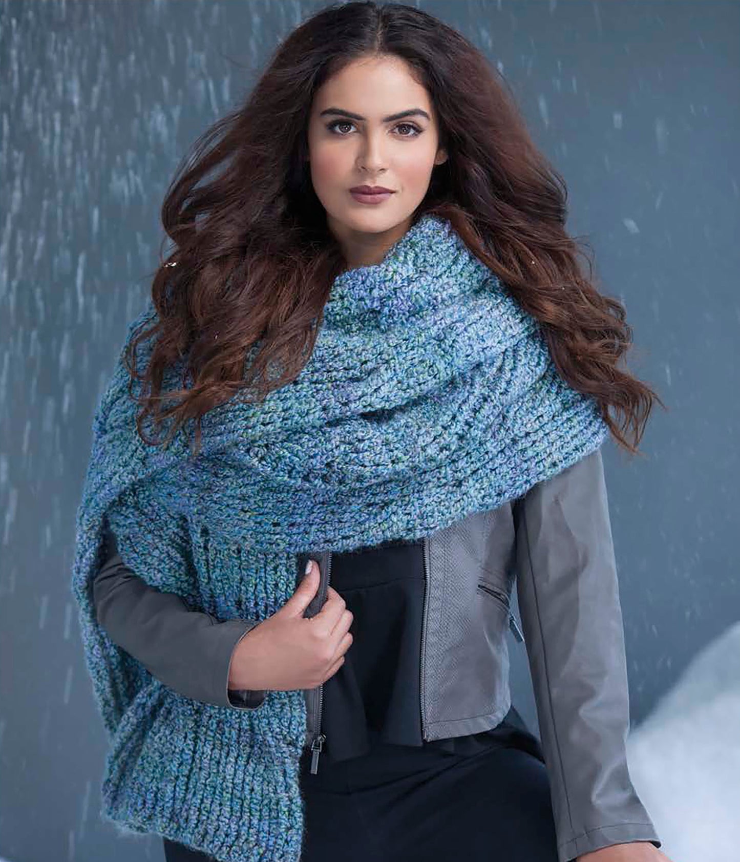 A woman with long wavy brown hair models a chunky blue scarf from Leisure Arts' Easy Super Scarves Crochet, draped over a gray jacket and standing before a snowy scene—perfect inspiration for craft book lovers and beginners alike.