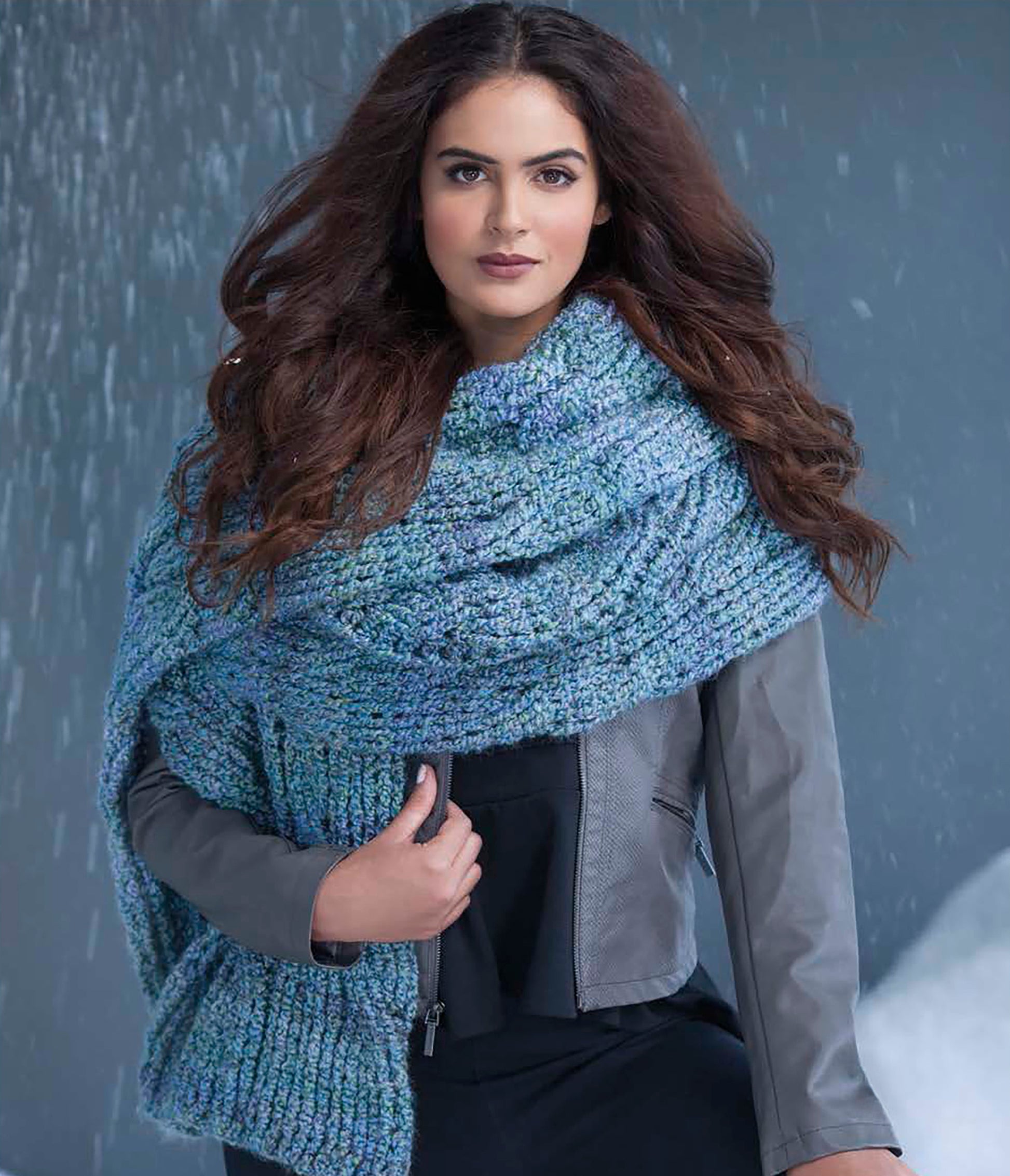 A woman with long wavy brown hair models a chunky blue scarf from Leisure Arts' Easy Super Scarves Crochet, draped over a gray jacket and standing before a snowy scene—perfect inspiration for craft book lovers and beginners alike.