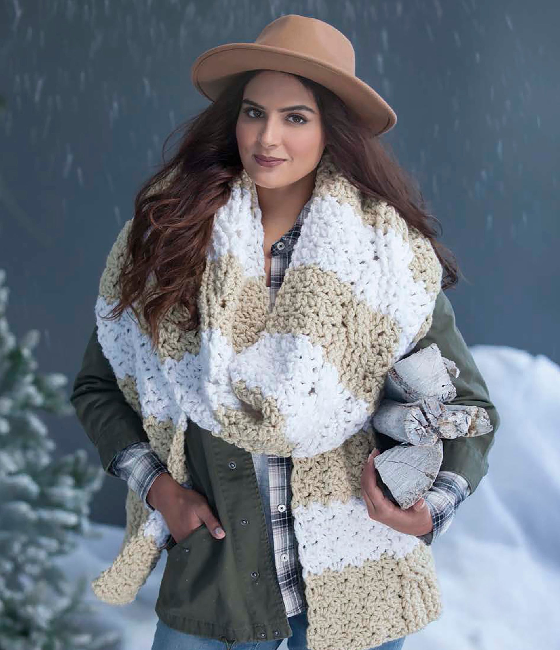 Wearing a tan hat, green jacket, and a large beige and white plaid scarf she crocheted with Easy Super Scarves Crochet by Leisure Arts, a woman stands smiling in snowy outdoors, holding rustic holiday decorations with pine trees behind her.
