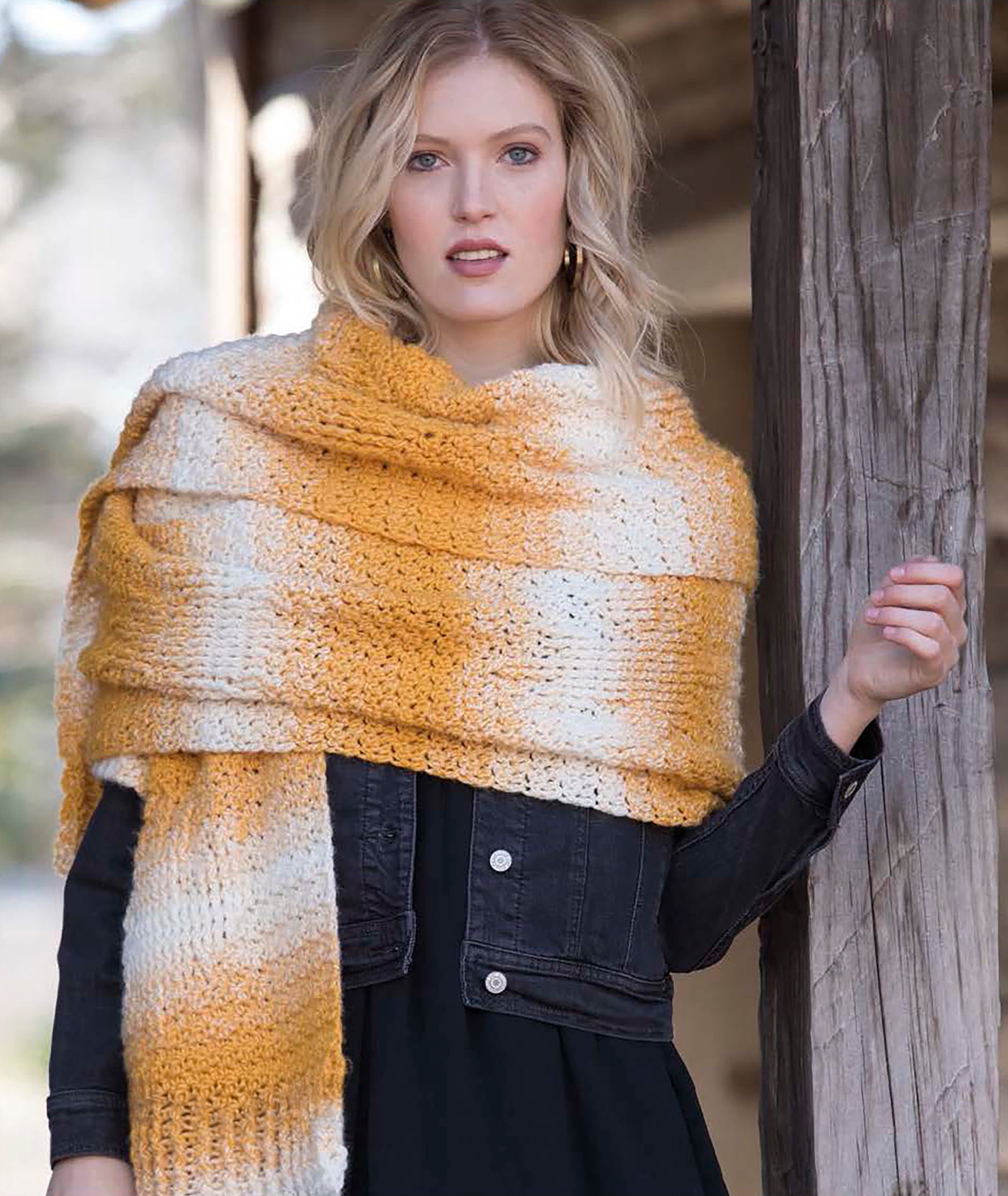 A blonde woman models a large yellow and white striped scarf, crocheted with the Easy Super Scarves Crochet pattern by Leisure Arts, over a black denim jacket and dress. She stands by a wooden post, looking at the camera.