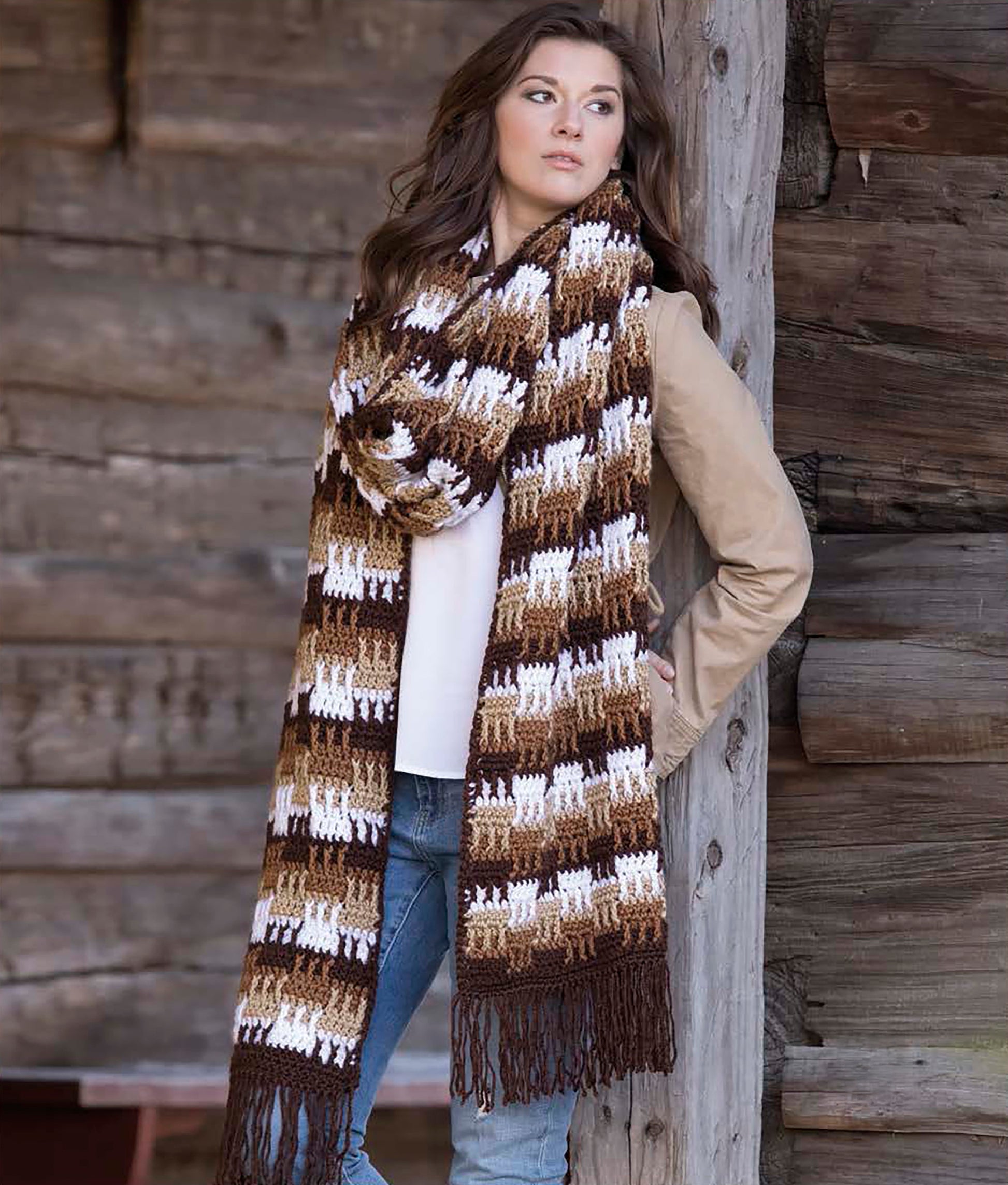 A woman leans against a wooden wall, modeling a large striped scarf made with the Easy Super Scarves Crochet book by Leisure Arts, styled with a beige jacket and blue jeans—perfect for beginner crocheters.