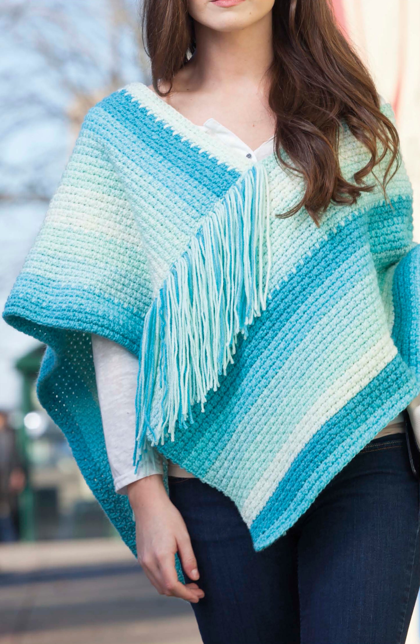 A woman stands outdoors wearing a blue and white striped crocheted poncho with fringe, made using Caron Cakes Crochet Projects by Leisure Arts, layered over a white top and paired with dark jeans.