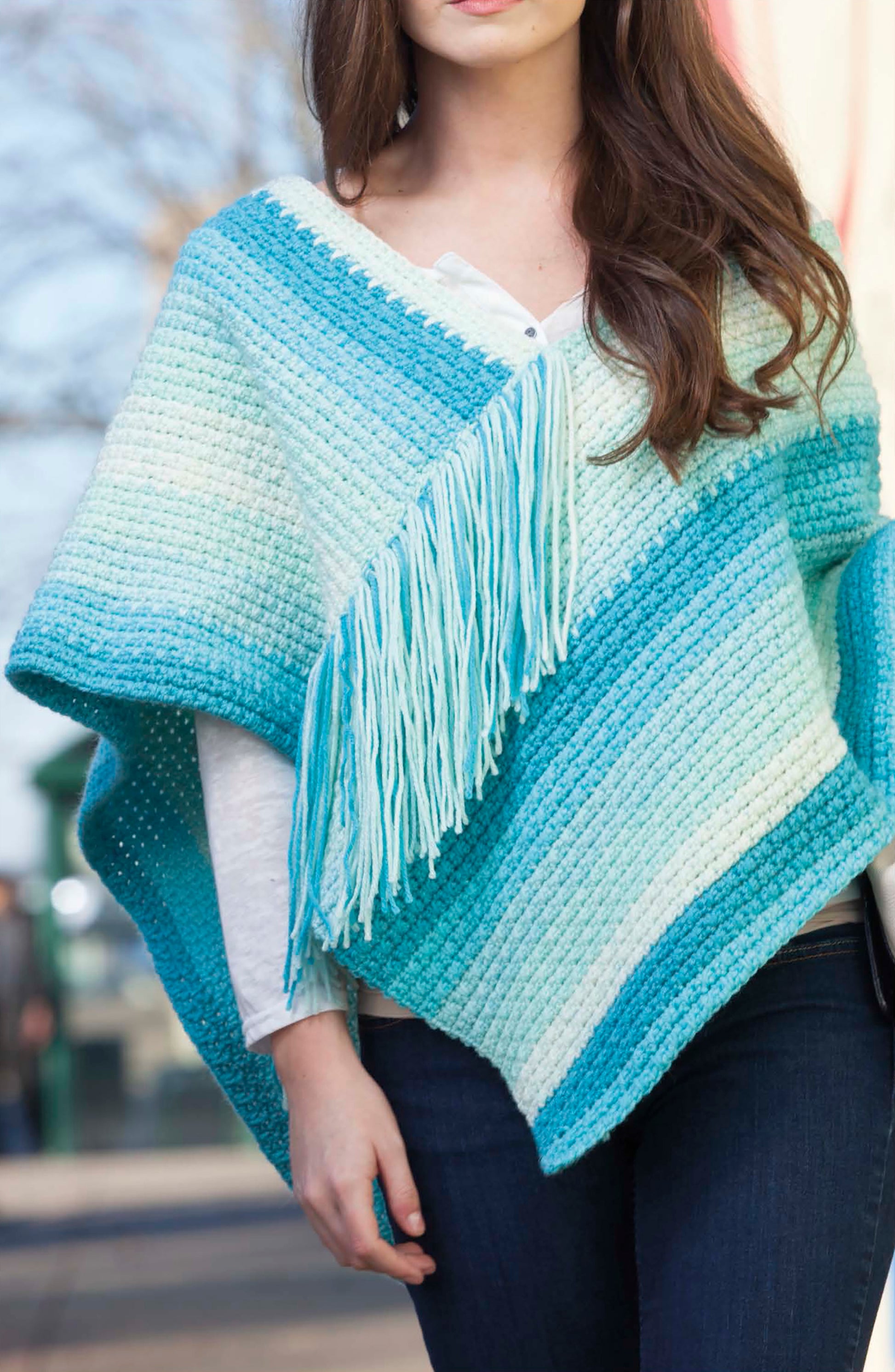 A woman stands outdoors wearing a blue and white striped crocheted poncho with fringe, made using Caron Cakes Crochet Projects by Leisure Arts, layered over a white top and paired with dark jeans.