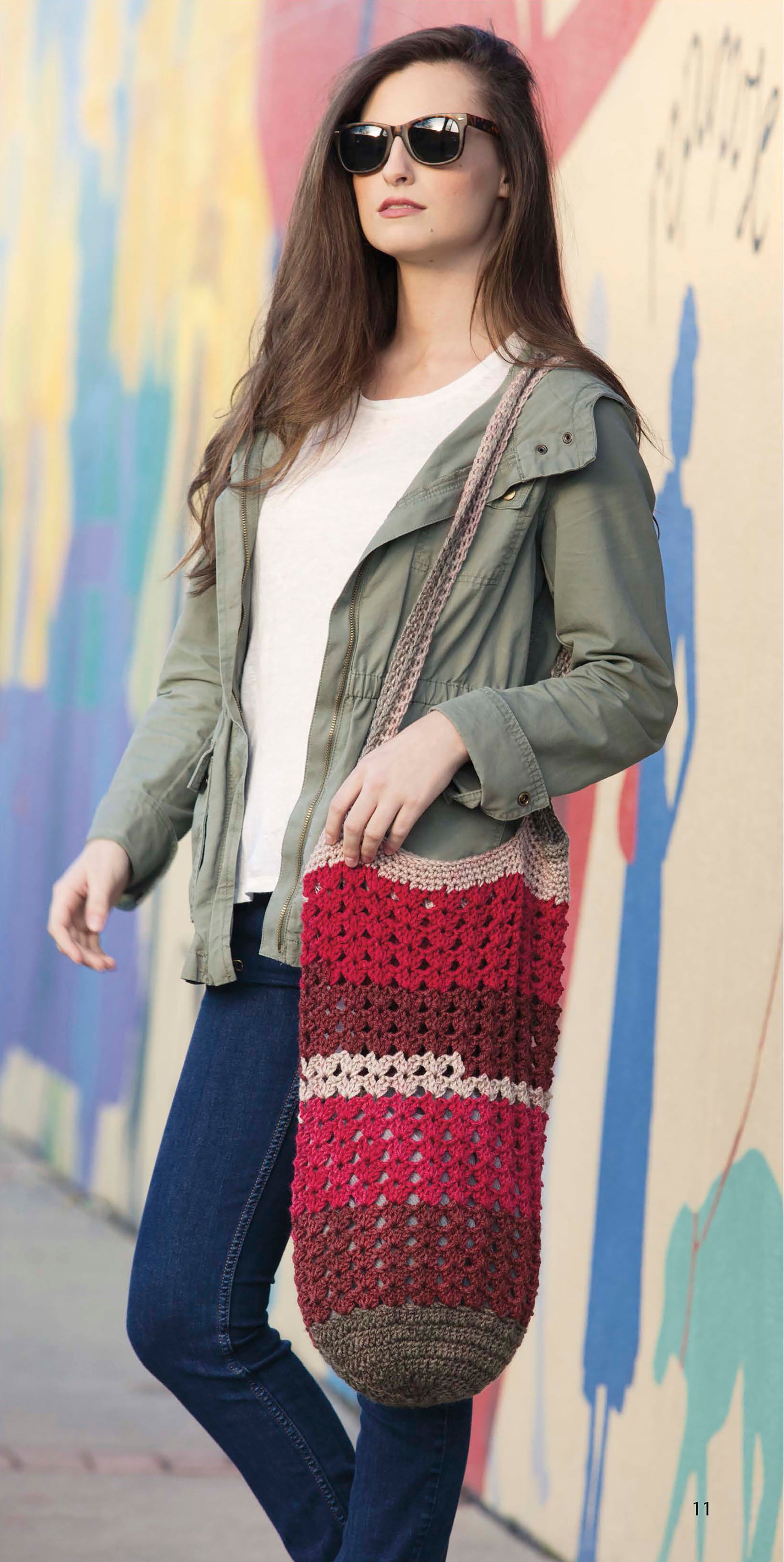 A woman with long brown hair, wearing sunglasses, a green jacket, white shirt, and jeans poses by a colorful mural holding a crocheted red, pink, and brown tote bag made using Leisure Arts’ Caron Cakes Crochet Projects.