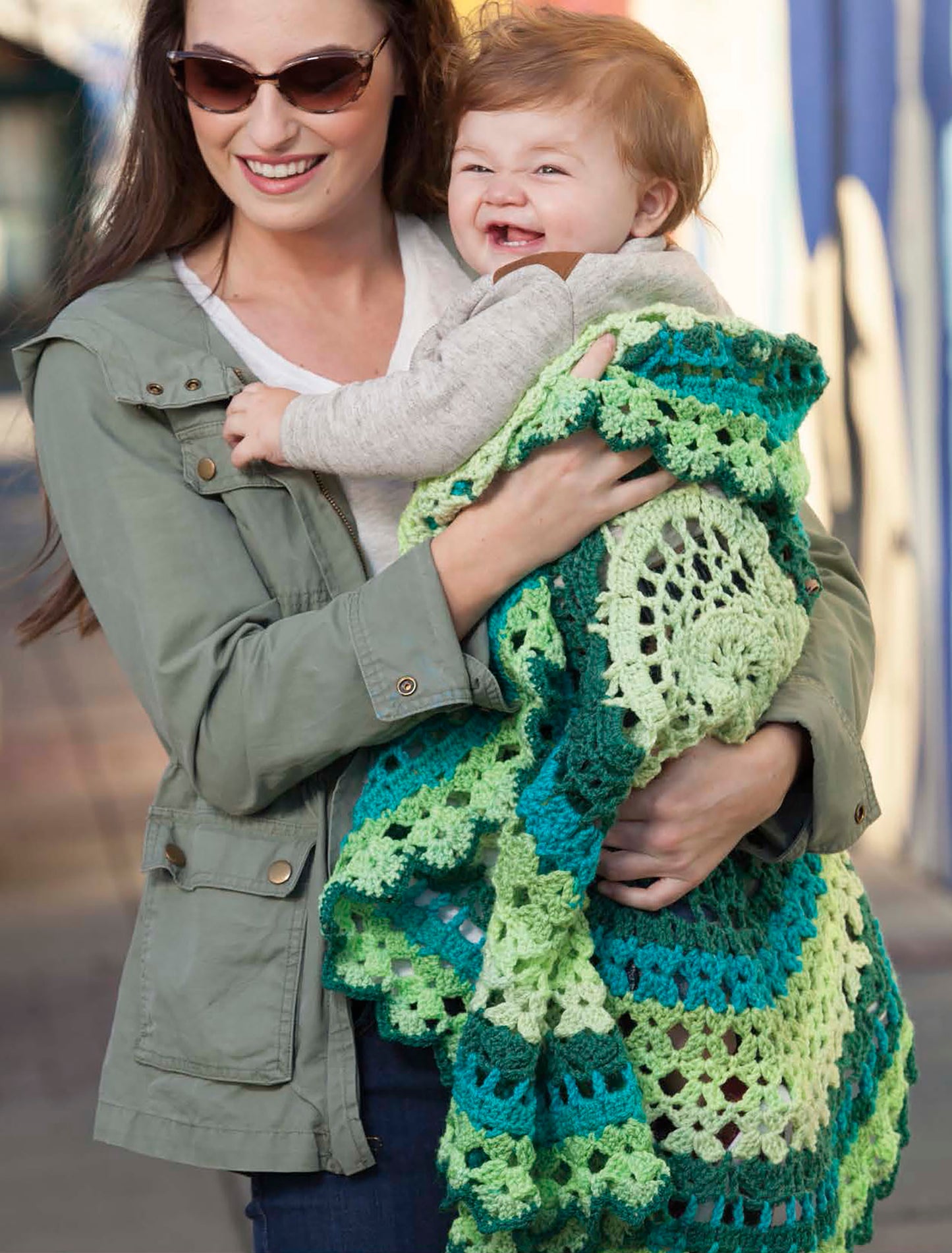 A woman in sunglasses holds a smiling baby wrapped in a green and blue crocheted blanket made with Caron Cakes, inspired by Leisure Arts’ crochet projects.