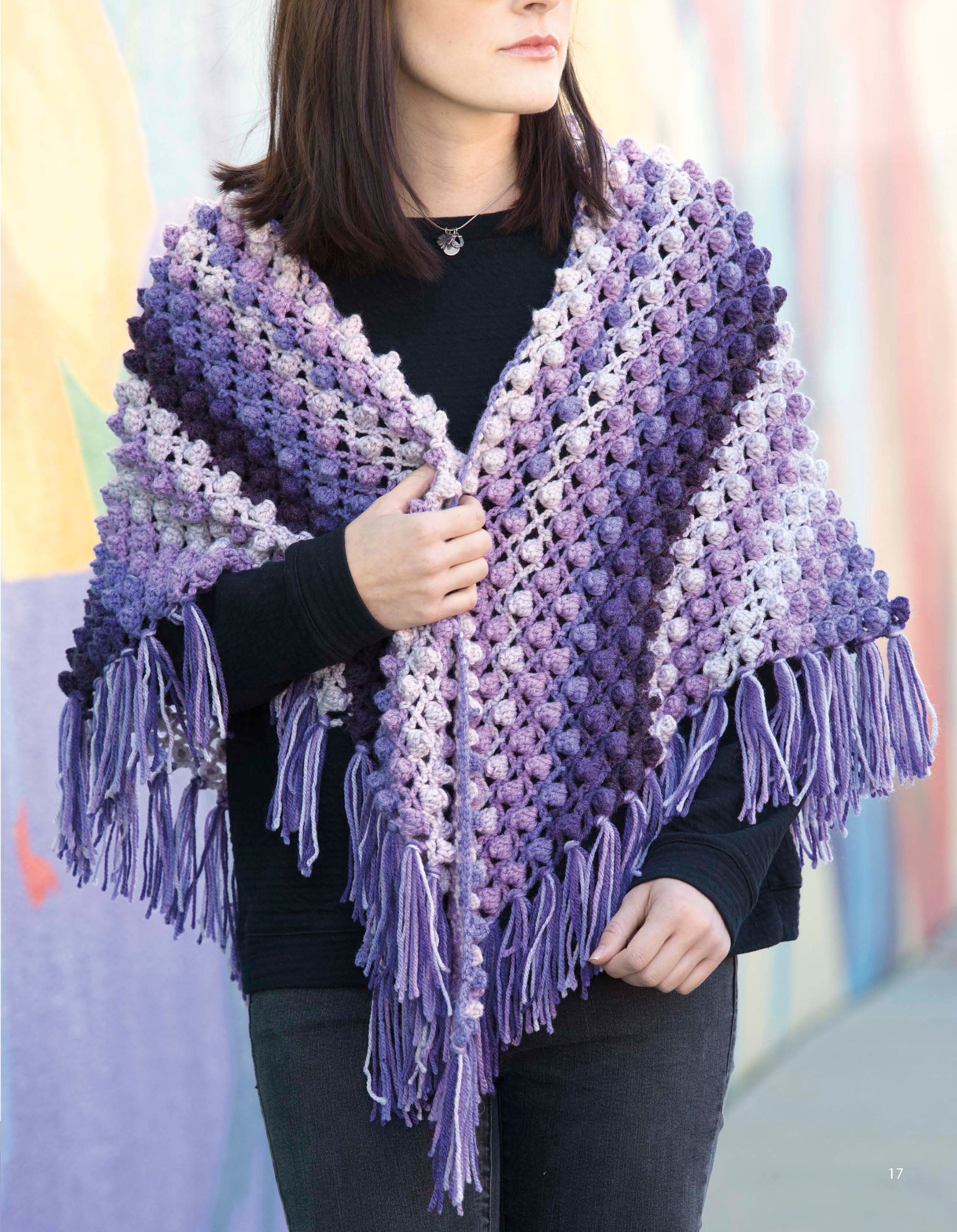 A woman in a black top and pants models a purple and lavender fringe shawl from "Caron Cakes Crochet Projects" by Leisure Arts, standing against a colorful wall and gently holding the cozy wrap around her shoulders.
