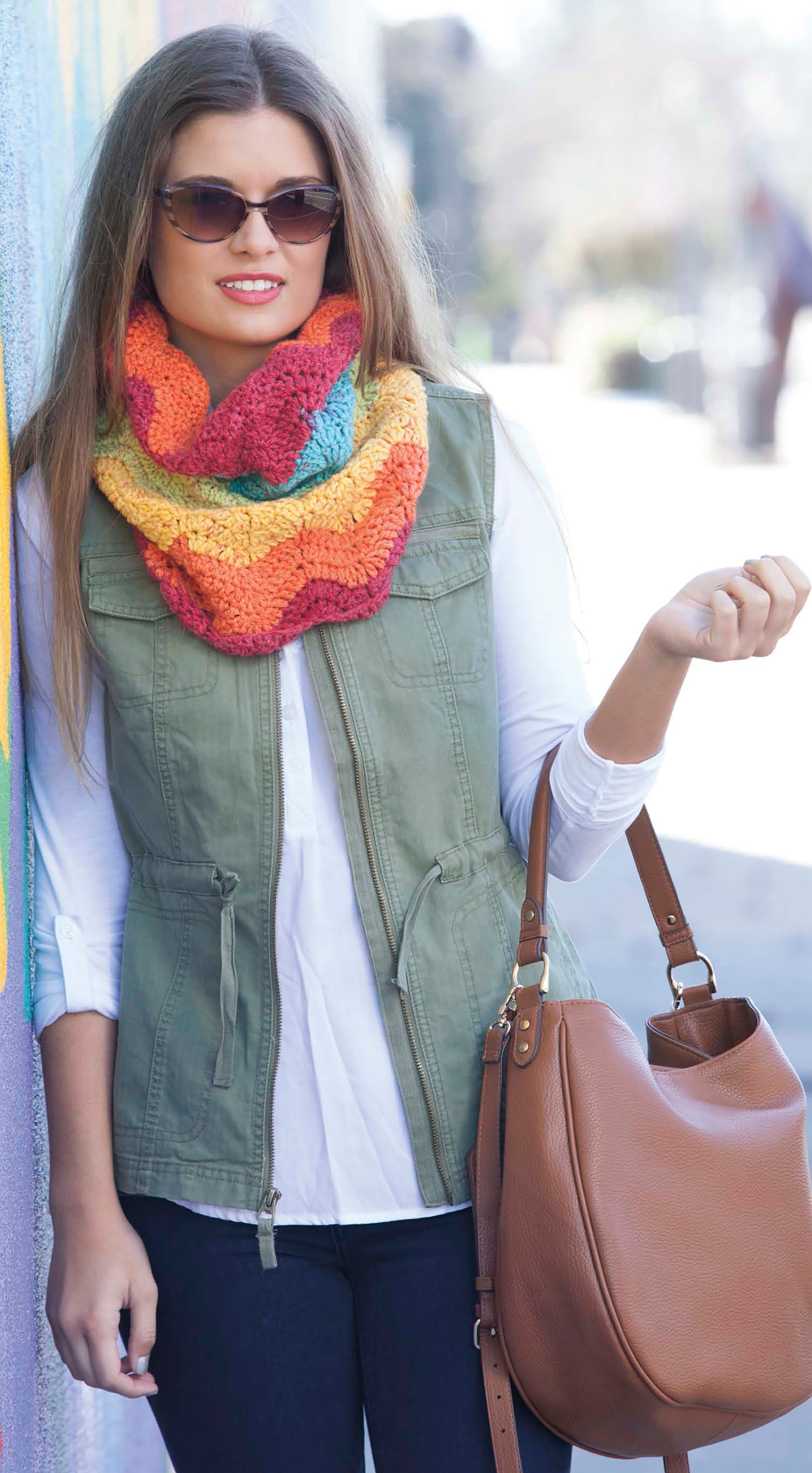 A woman wearing sunglasses, a green utility vest over a white shirt, dark jeans, and a multicolored knit scarf made with Caron Cakes Crochet Projects by Leisure Arts holds a large tan handbag while standing against a wall outdoors.
