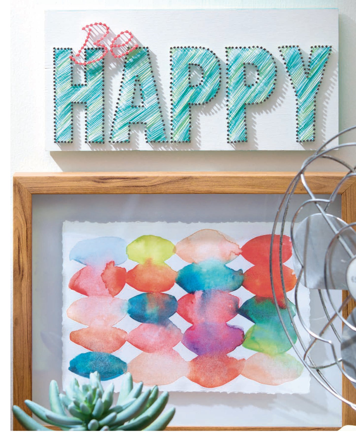 The Cool String Art by Leisure Arts, featuring a "Be Happy" design with precise nail placement, is displayed above a framed abstract watercolor painting, with a succulent plant and part of a metal fan in the foreground.