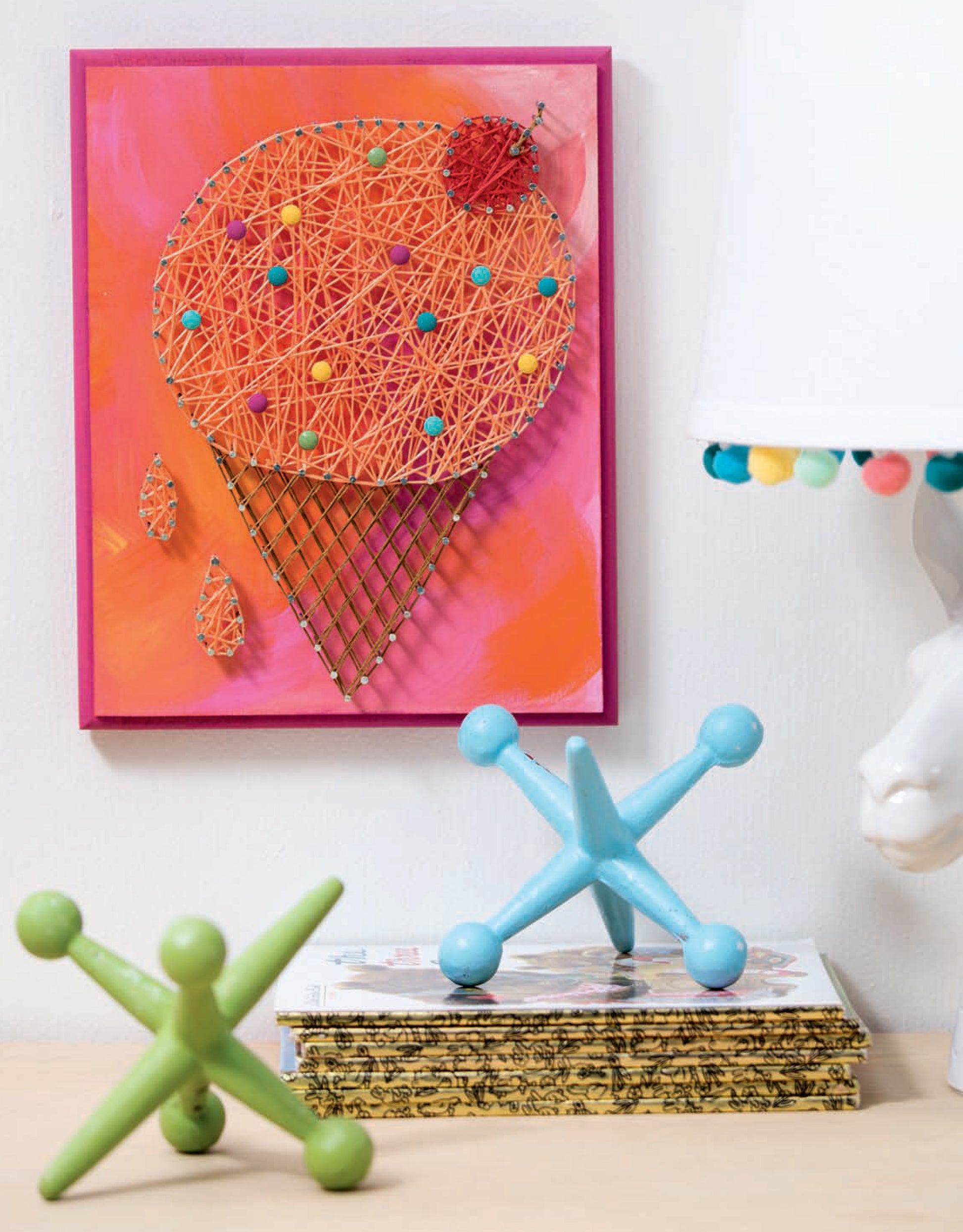 The Cool String Art by Leisure Arts, a vibrant wall decor shaped like an ice cream cone, features a unique nail pattern and hangs above a table with decorative jacks, stacked books, and a white lamp with pompom-trimmed shade.