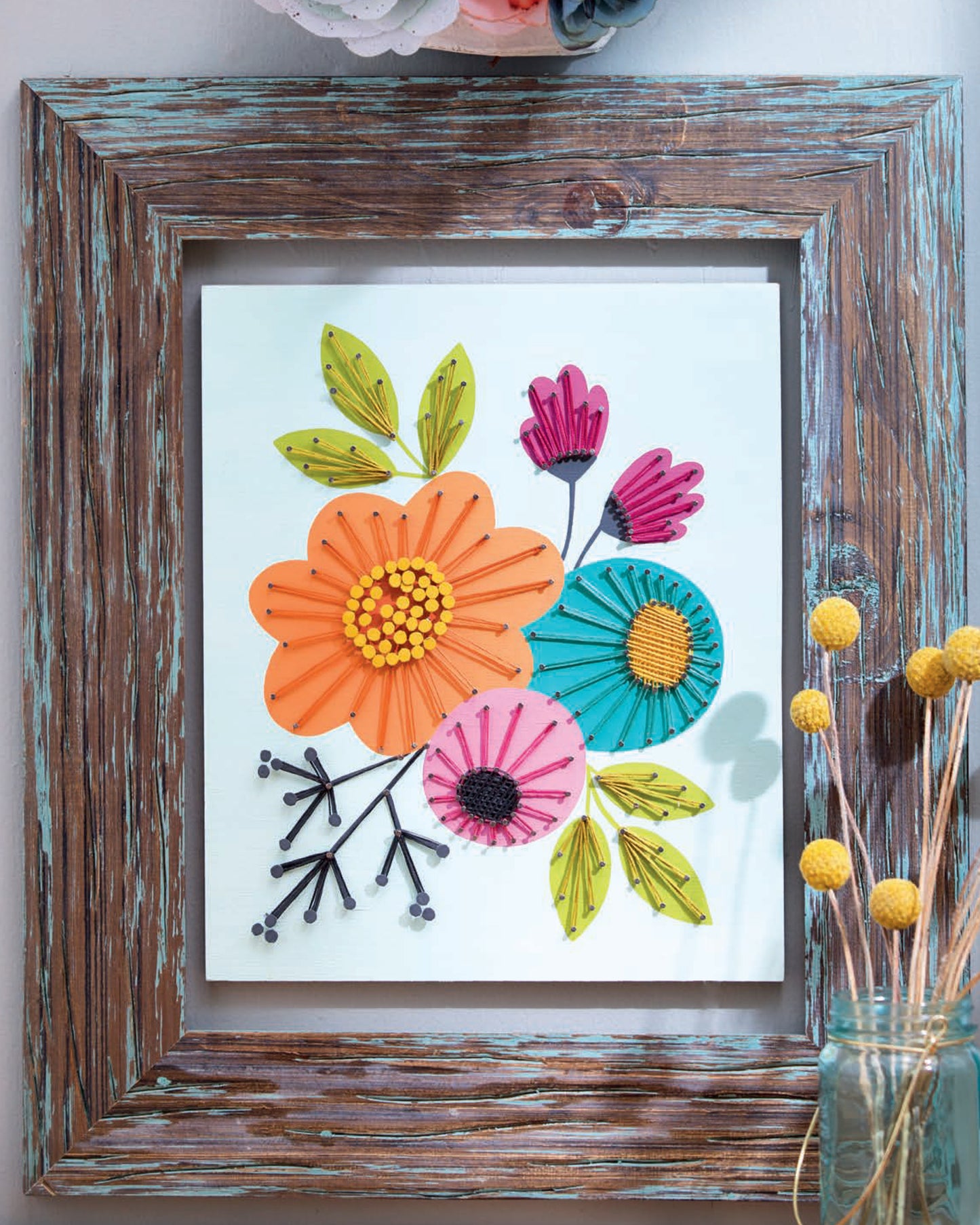 Cool String Art by Leisure Arts is a framed artwork showcasing three-dimensional, colorful paper flowers and leaves on a white background, with a jar of yellow billy balls in the foreground amid a rustic wooden frame.