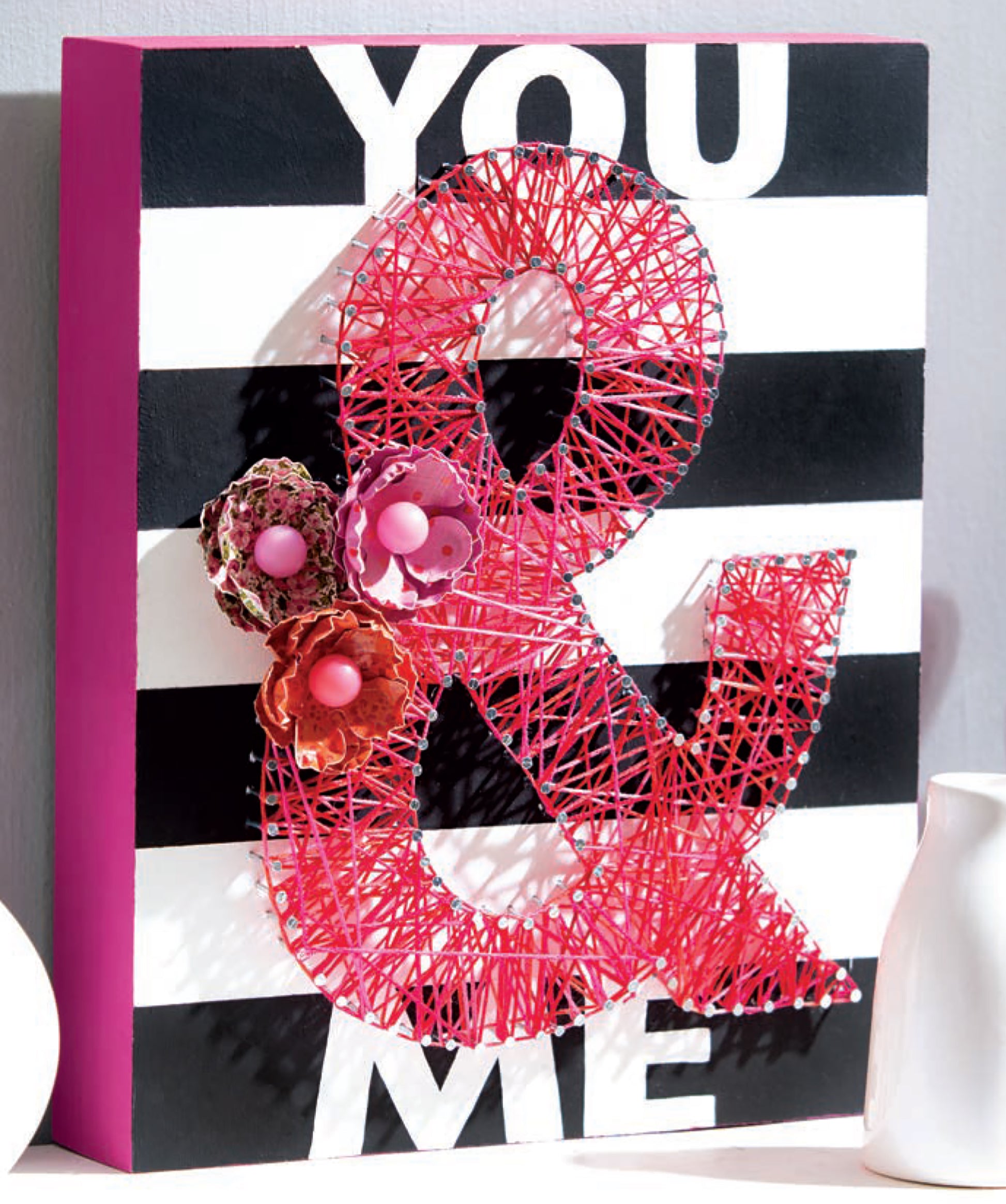 The Cool String Art by Leisure Arts is a decorative black and white striped sign with "YOU & ME" text. The red string art ampersand features three handcrafted, colorful flower embellishments for a charming touch.