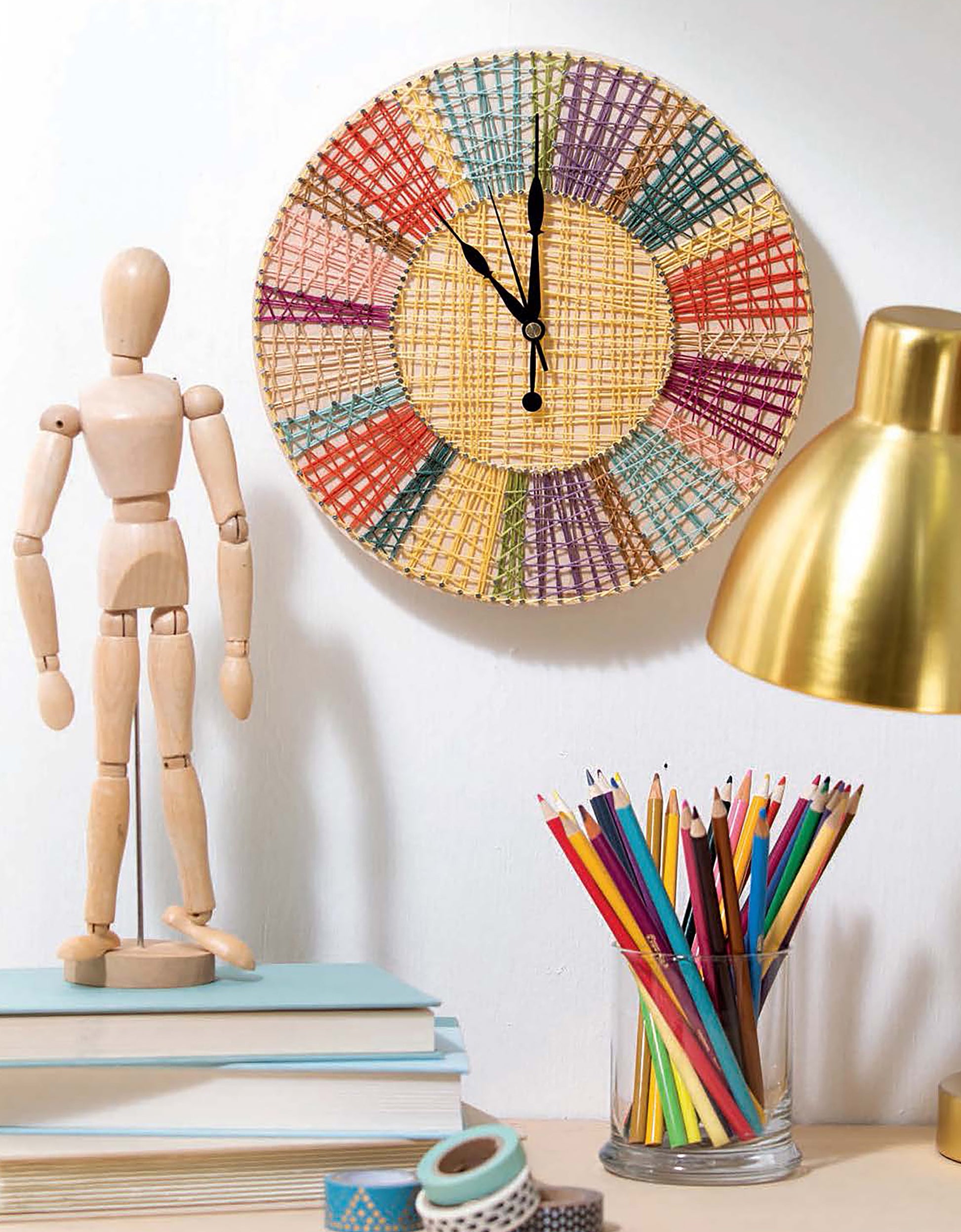 A Cool String Art wall clock from Leisure Arts marks 11:00, with a wooden mannequin on books, gold lamp, and glass jar of colored pencils nearby—ideal for sparking craft ideas or DIY panel projects at your desk.