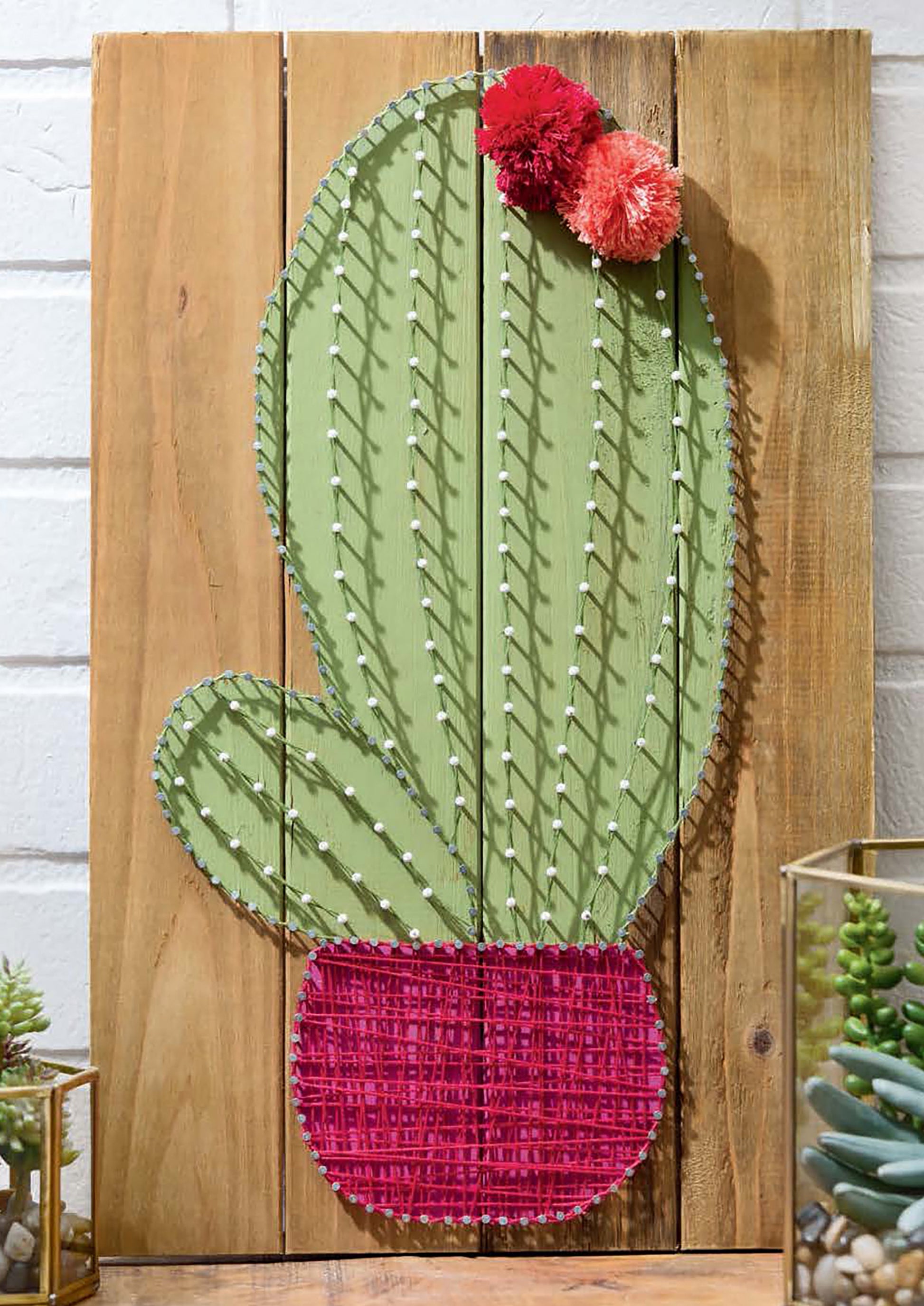 Cool String Art by Leisure Arts features a cactus outlined in green string on wooden boards, a pink string pot, and two pom-pom flowers. Succulents and décor accents are included nearby for added style.