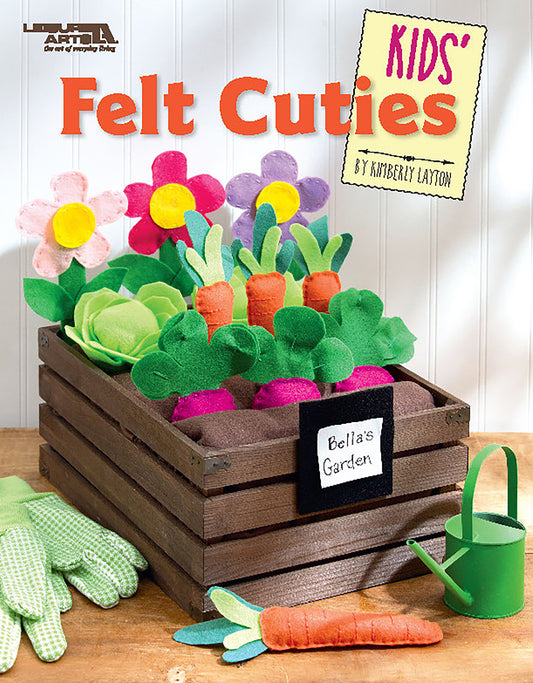 A wooden crate labeled Leisure Arts Kids' Felt Cuties holds colorful felt veggies and flowers, perfect for kids’ crafts. Green gardening gloves and a small watering can are arranged nearby on a white surface.