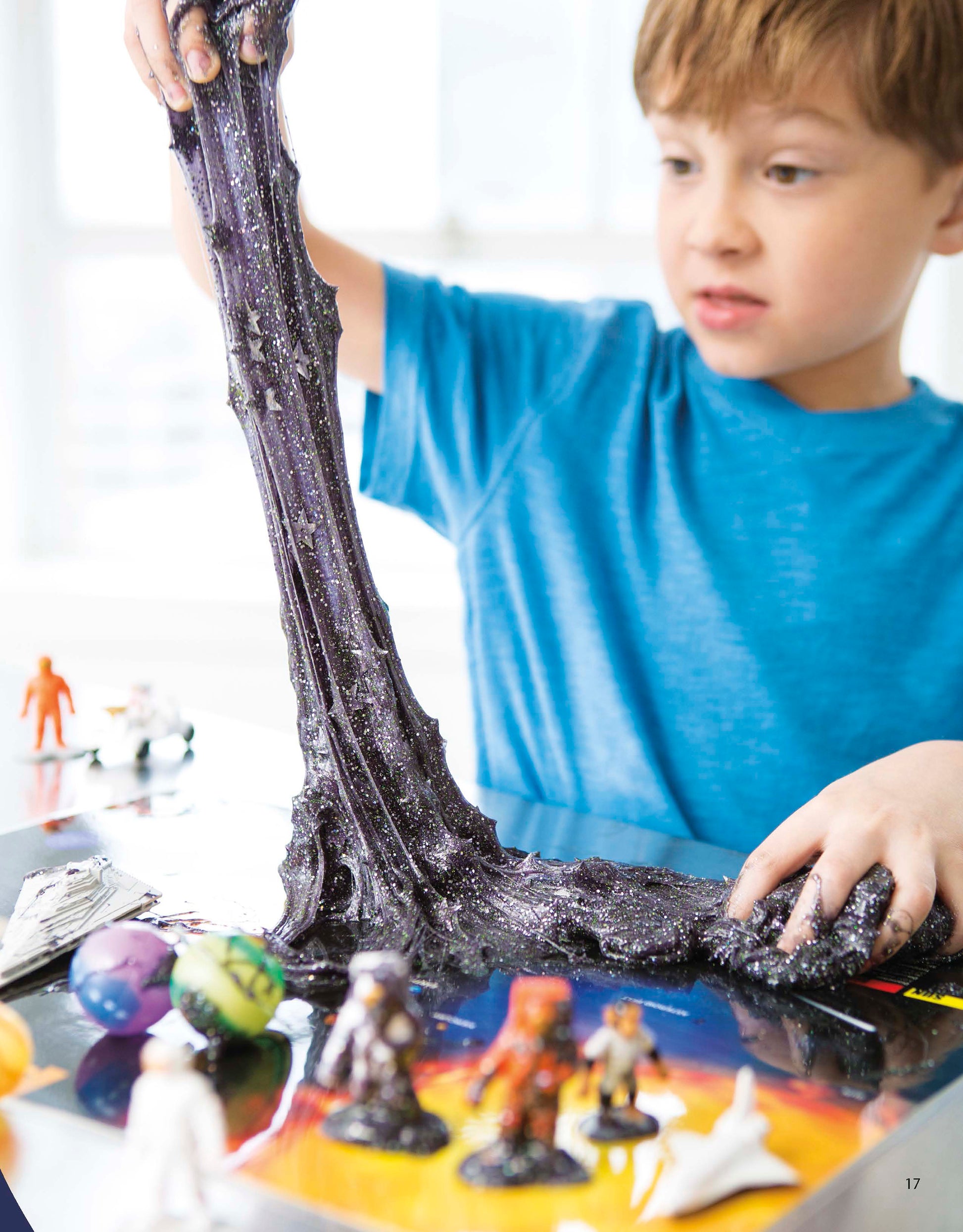 A young boy in a blue shirt stretches shiny black slime with star-shaped glitter, surrounded by toys—a fun craft inspired by Slimed DIY—A guide to making Slime at home! Digital Download from Leisure Arts. Perfect for creative kids.