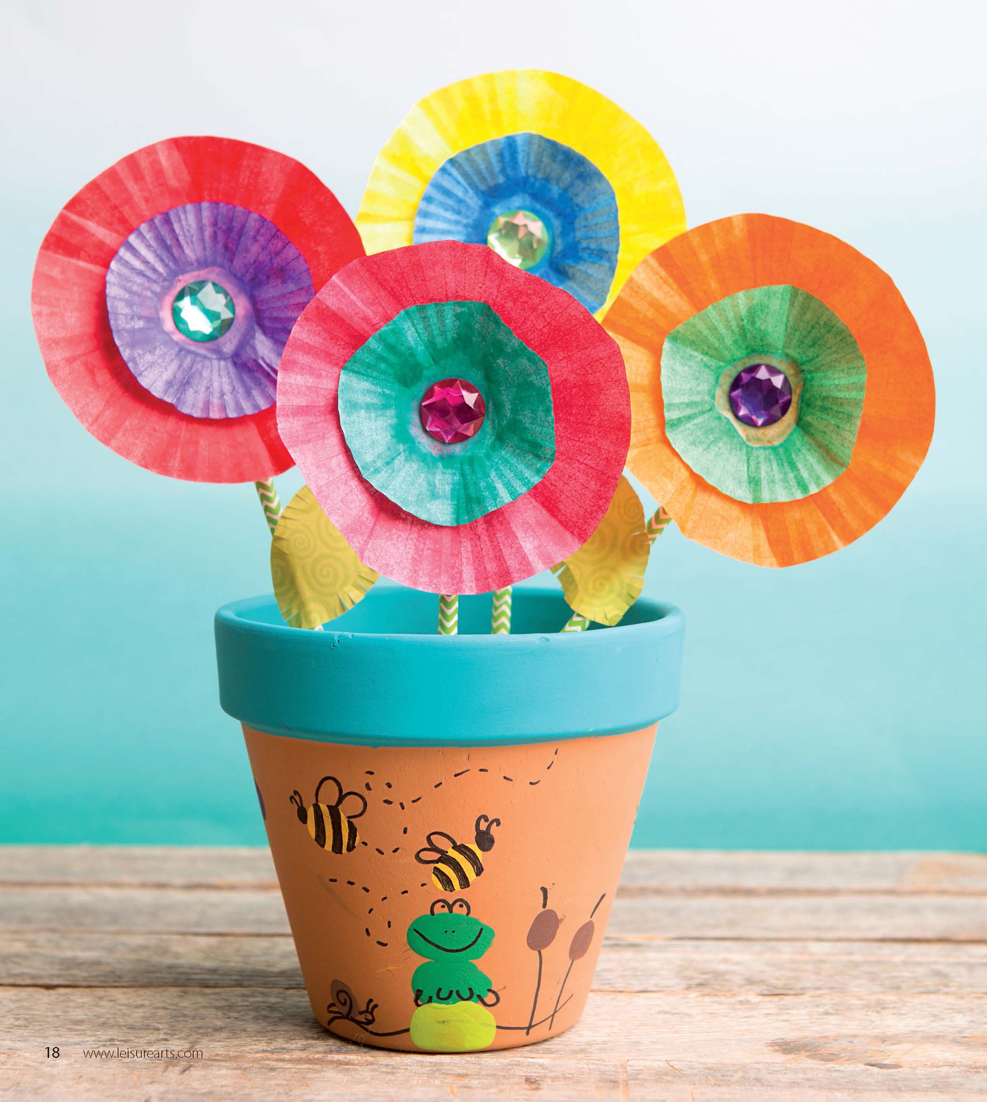The Leisure Arts DIY Simple Kids' Crafts set features a painted flowerpot with a frog and bees holding colorful paper flowers with gemstone centers, perfect for kids’ art projects, displayed against a blue background.