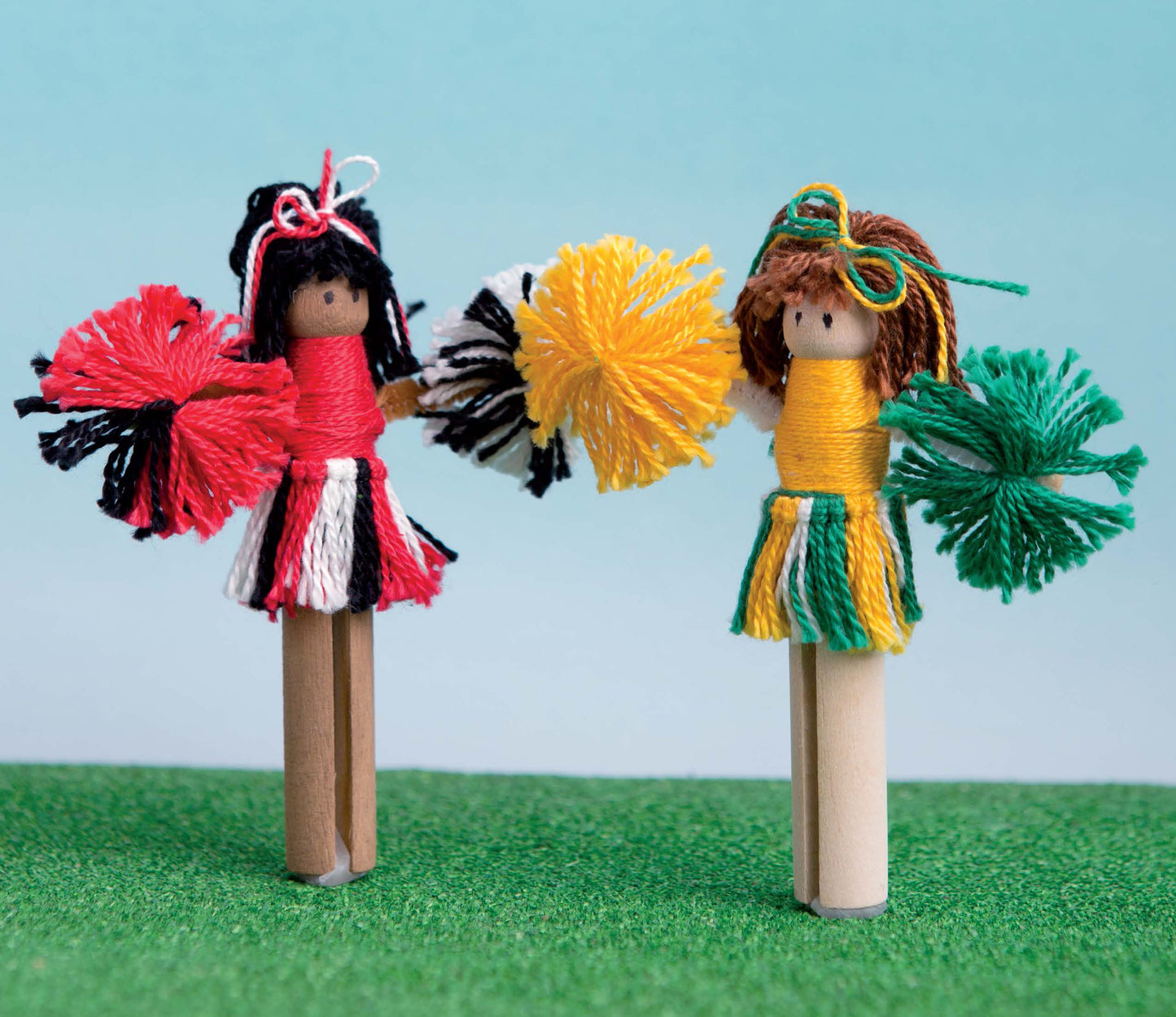 Two handmade cheerleader dolls, crafted from clothespins and yarn pom-poms, stand on grass in bright uniforms. Perfect for DIY Simple Kids' Crafts by Leisure Arts—great for craft kit fun or kids’ art projects with a sky blue background.