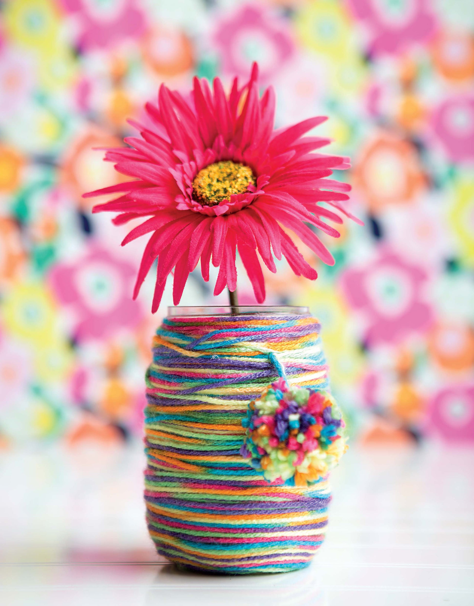 Create cheerful kids’ art projects with Leisure Arts’ DIY Simple Kids' Crafts. This craft features a bright pink flower in a yarn-wrapped vase topped with a pom-pom, set against a vivid, floral background—fun and easy for little crafters!.