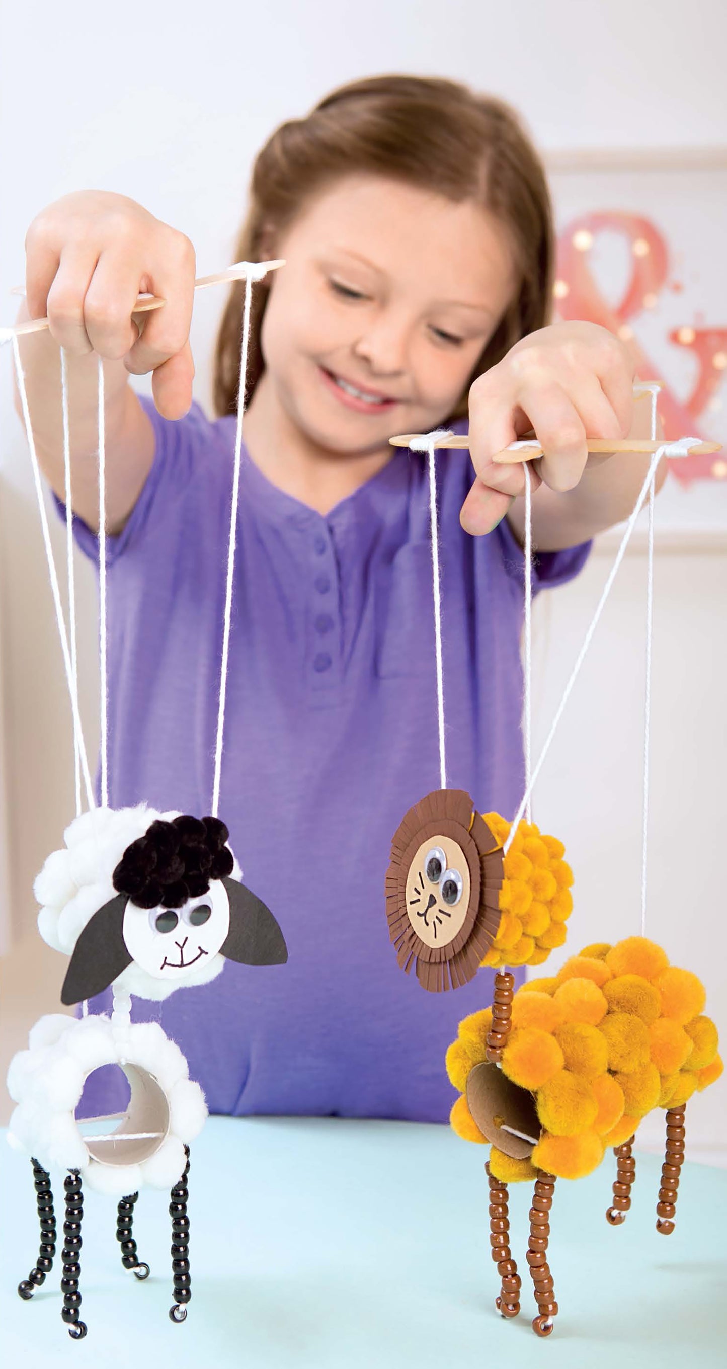 A smiling girl in a purple shirt holds two animal marionettes—a sheep and a lion—made with the DIY Simple Kids' Crafts kit from Leisure Arts, perfect for kids’ art projects using pom-poms, pipe cleaners, and cardboard.