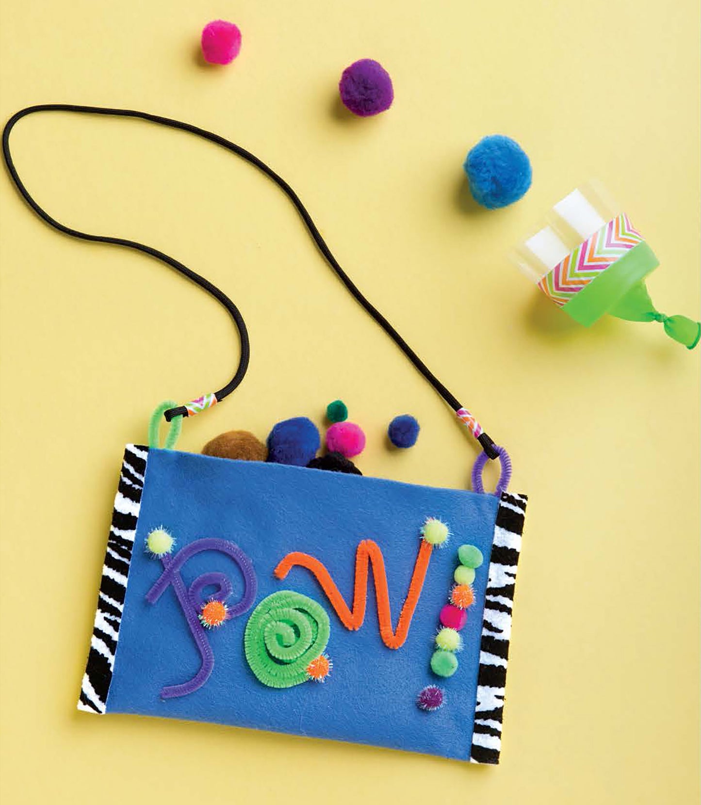 The DIY Simple Kids' Crafts bag by Leisure Arts features a blue felt front with pom-poms and pipe cleaner shapes, black strap, zebra-patterned sides, and is shown on a yellow background with pom-poms and a green cup—perfect for kids' creativity.