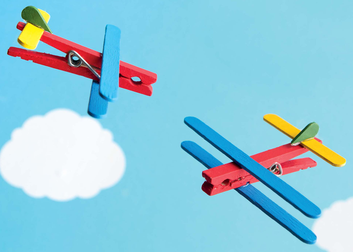 Two vibrant toy airplanes made from clothespins and popsicle sticks fly against a blue sky, inspiring creative activities for children. Discover more fun projects in DIY Simple Kids' Crafts by Leisure Arts.