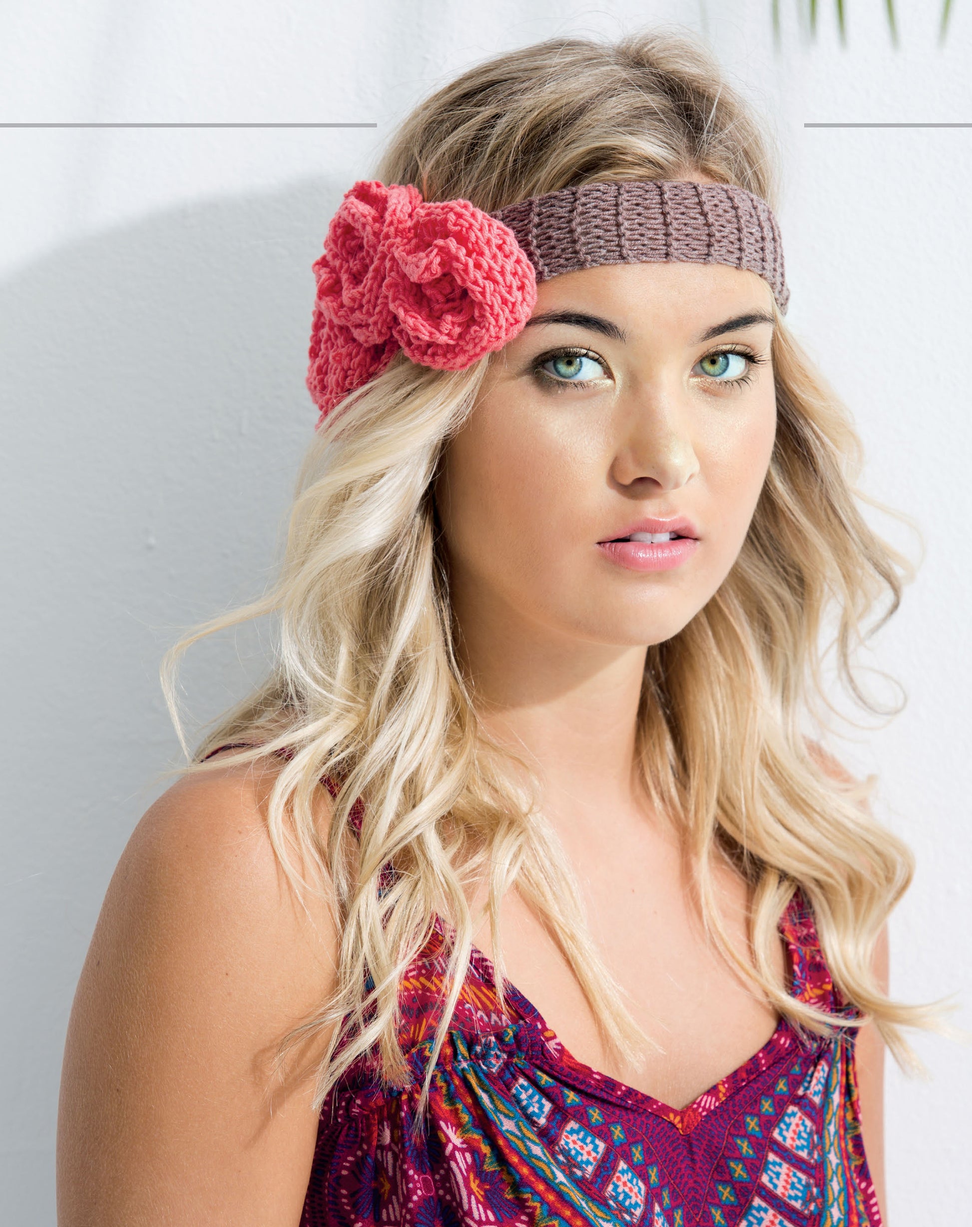 A young woman with long blonde hair wears a brown knitted headband with a large pink flower made from the Knits For Summer Digital Download by Leisure Arts, paired with a colorful top, posing against a light background.