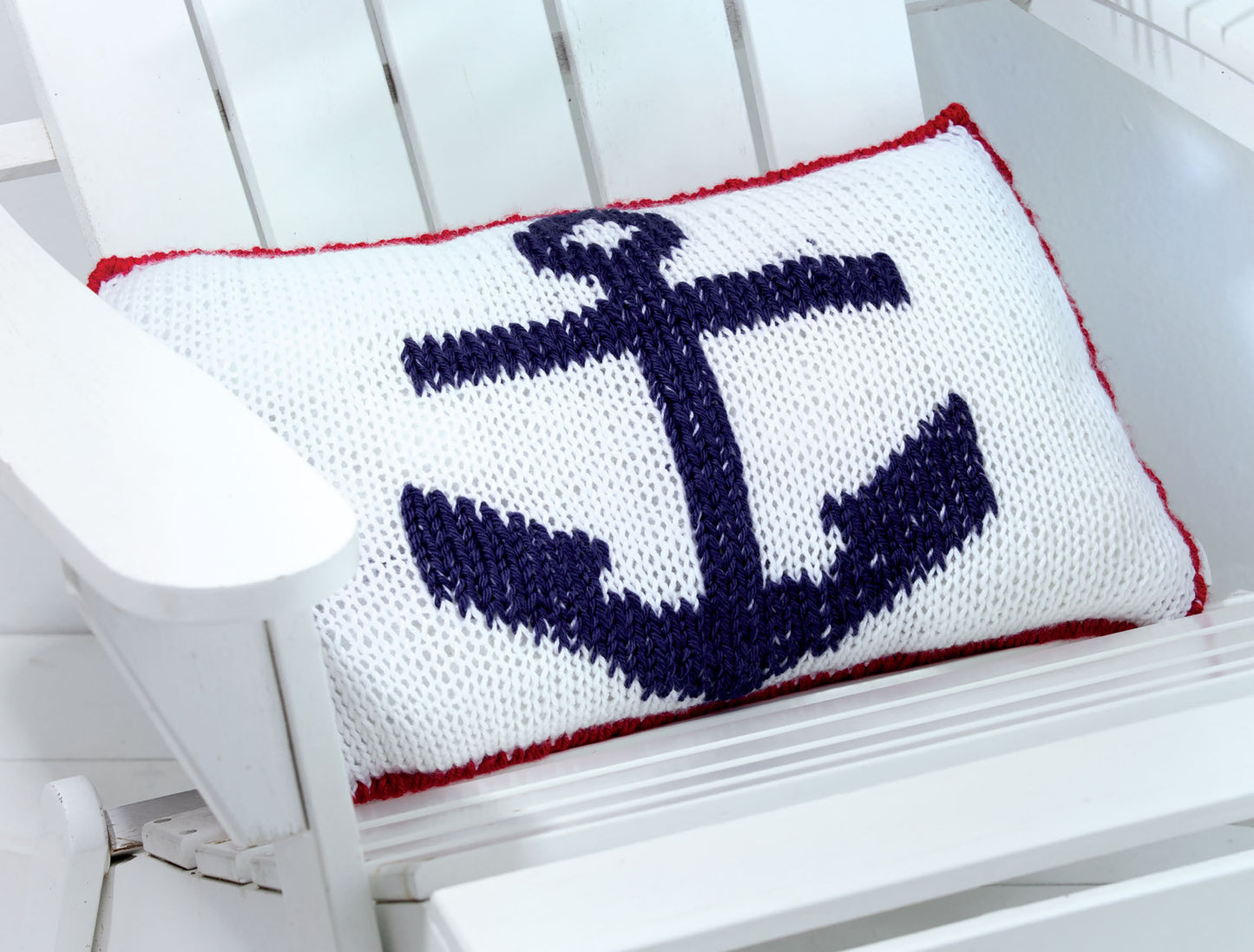 A white knitted cushion with a navy anchor and red trim, inspired by Knits For Summer Digital Download from Leisure Arts, sits on a white chair—perfect for fans of summer knit accessories.