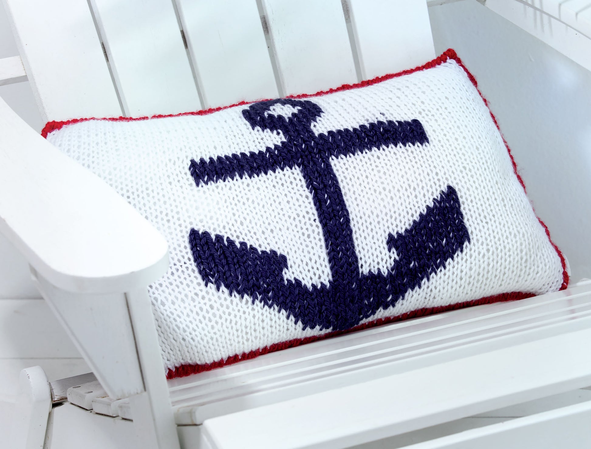 A white knitted cushion with a navy anchor and red trim, inspired by Knits For Summer Digital Download from Leisure Arts, sits on a white chair—perfect for fans of summer knit accessories.
