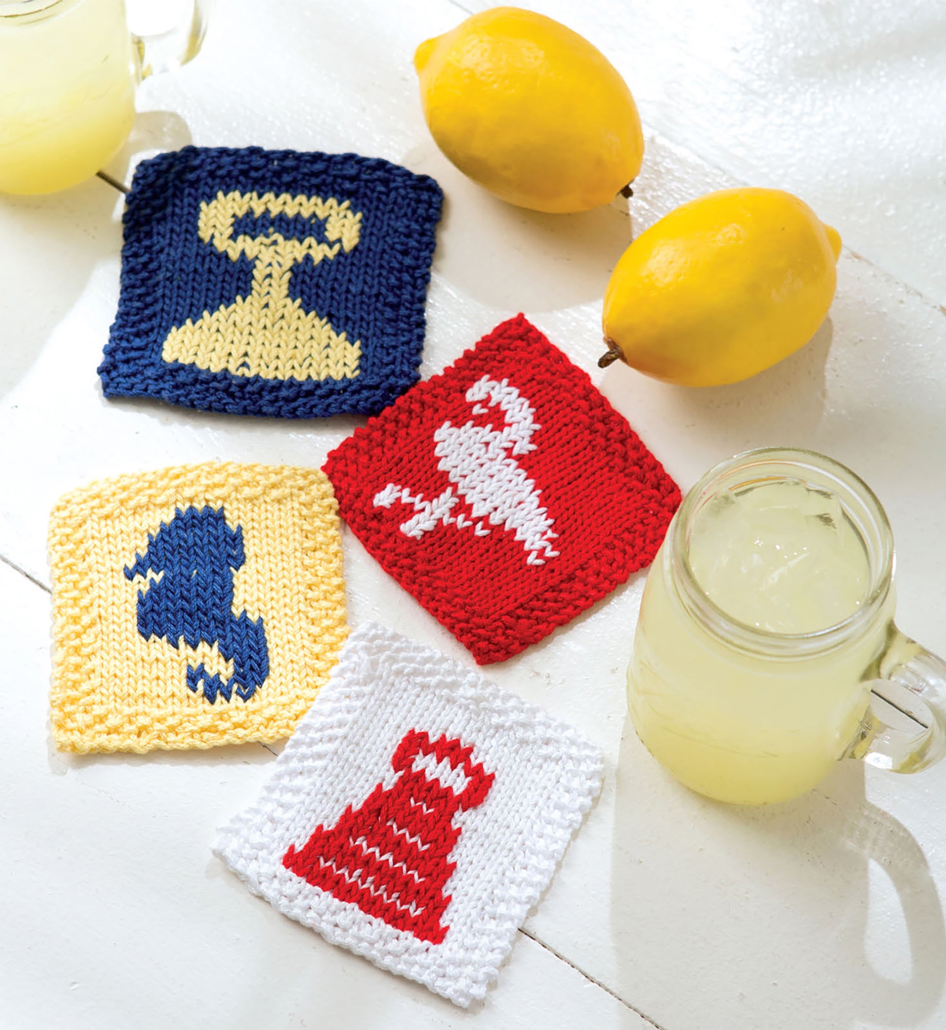 Leisure Arts' Knits For Summer Digital Download features four cotton coasters with trophy and pitcher designs in yellow, blue, red, and white, displayed on a white surface with two glasses of lemonade and two whole lemons.