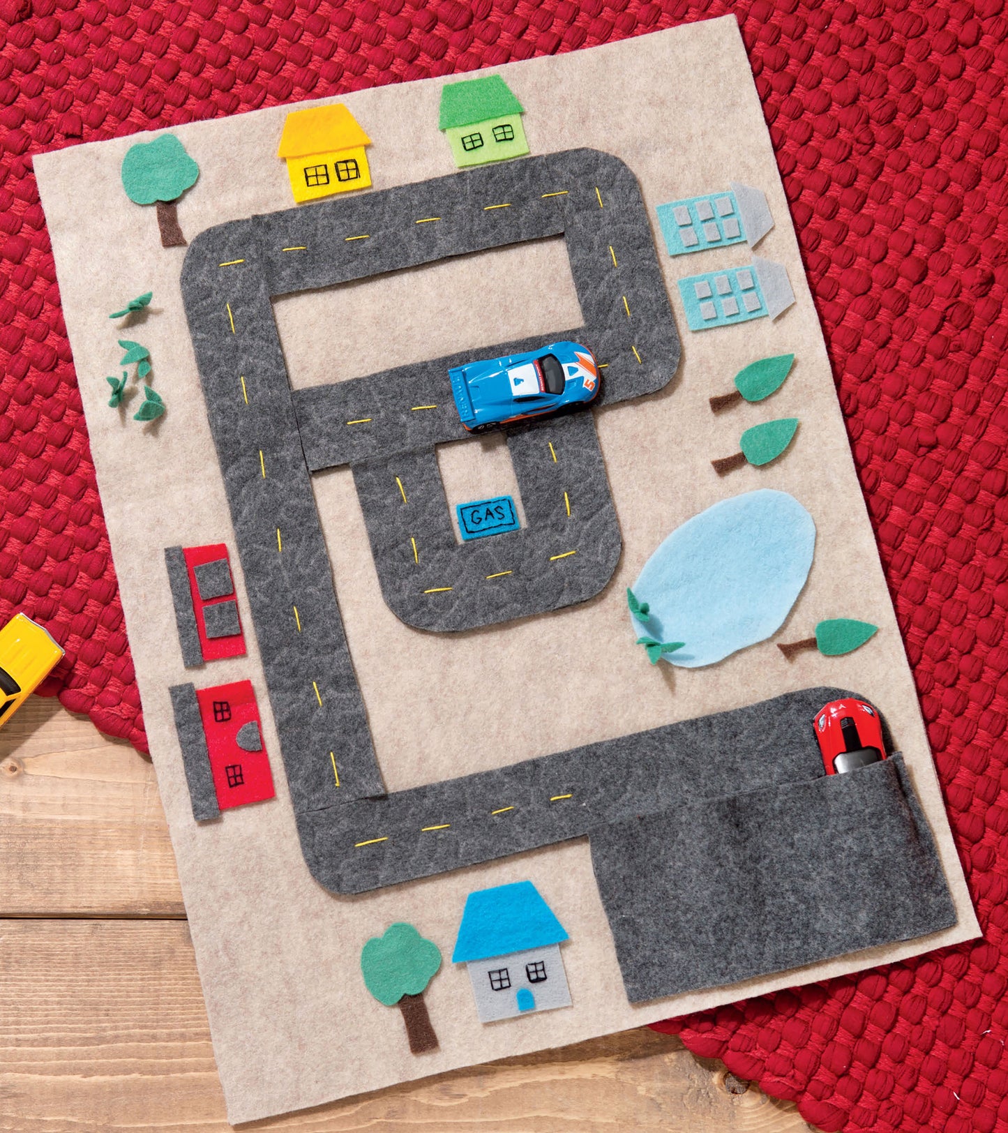 Leisure Arts’ Play Mats—7 Interactive Felt Designs for Making Your Own Story features a felt mat with roads, houses, trees, toy cars, a gas station, and a cupcake shop on a wooden surface with a red placemat.