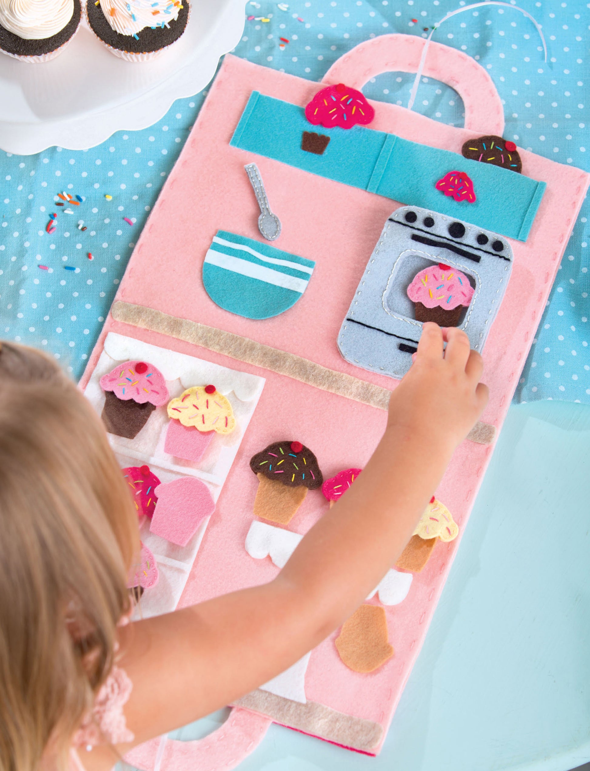 A young child enjoys the Leisure Arts Play Mats—7 Interactive Felt Designs for Making Your Own Story, using felt pieces shaped like kitchen items and cupcakes to create imaginative scenes inspired by magical treats, while real cupcakes rest on a cake stand.